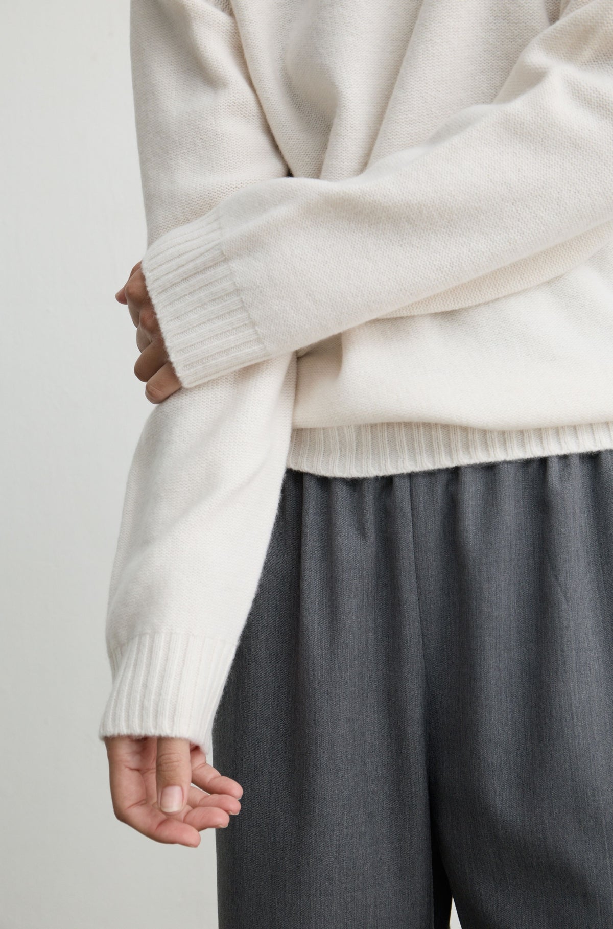 Close-up of a person wearing a cream knit sweater with ribbed cuffs and hem, paired with dark gray wide-leg trousers, against a plain light background.