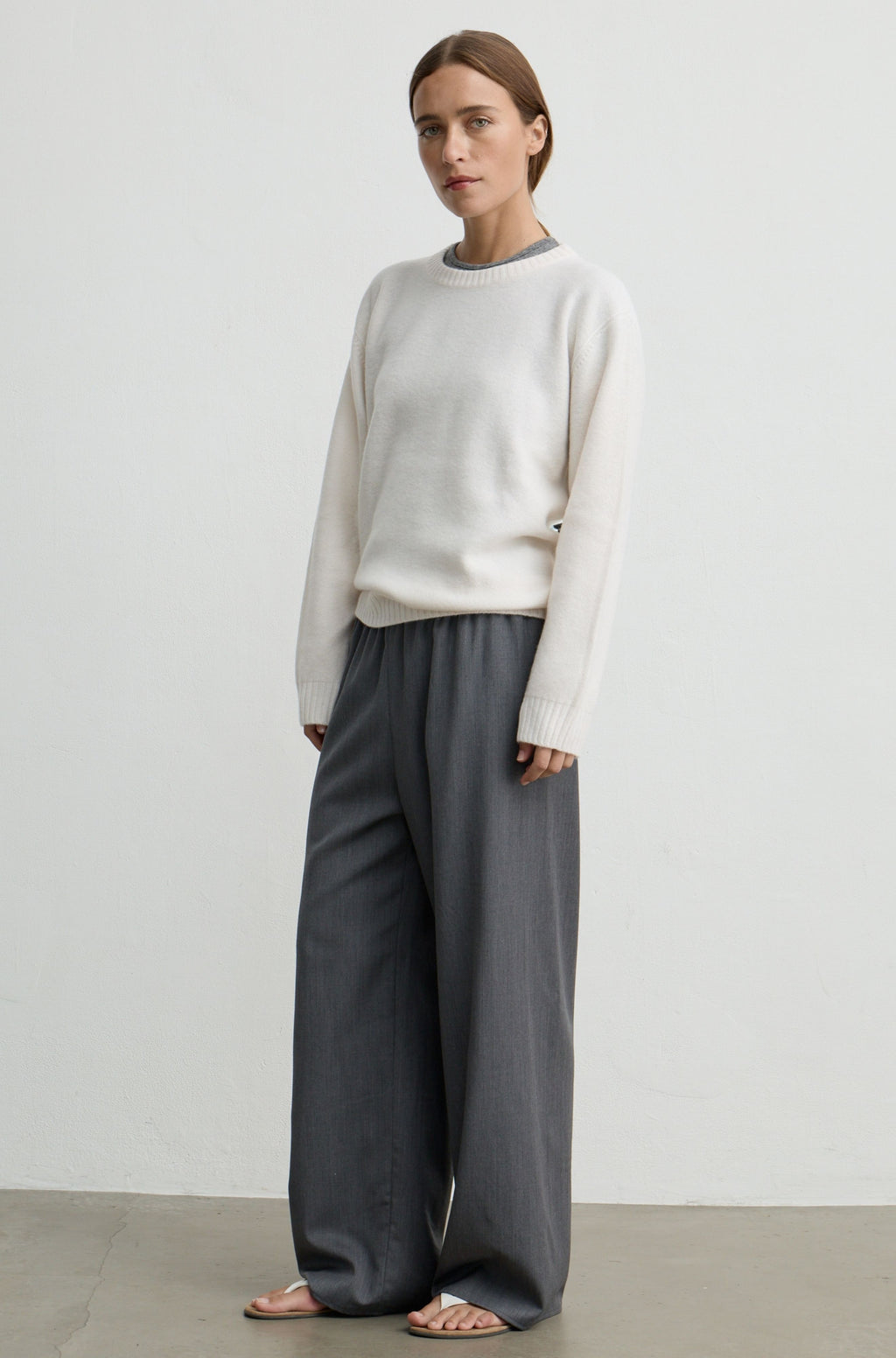 A woman modeling a cream knit crewneck sweater and dark gray wide-leg trousers, standing against a minimalist off-white studio wall.