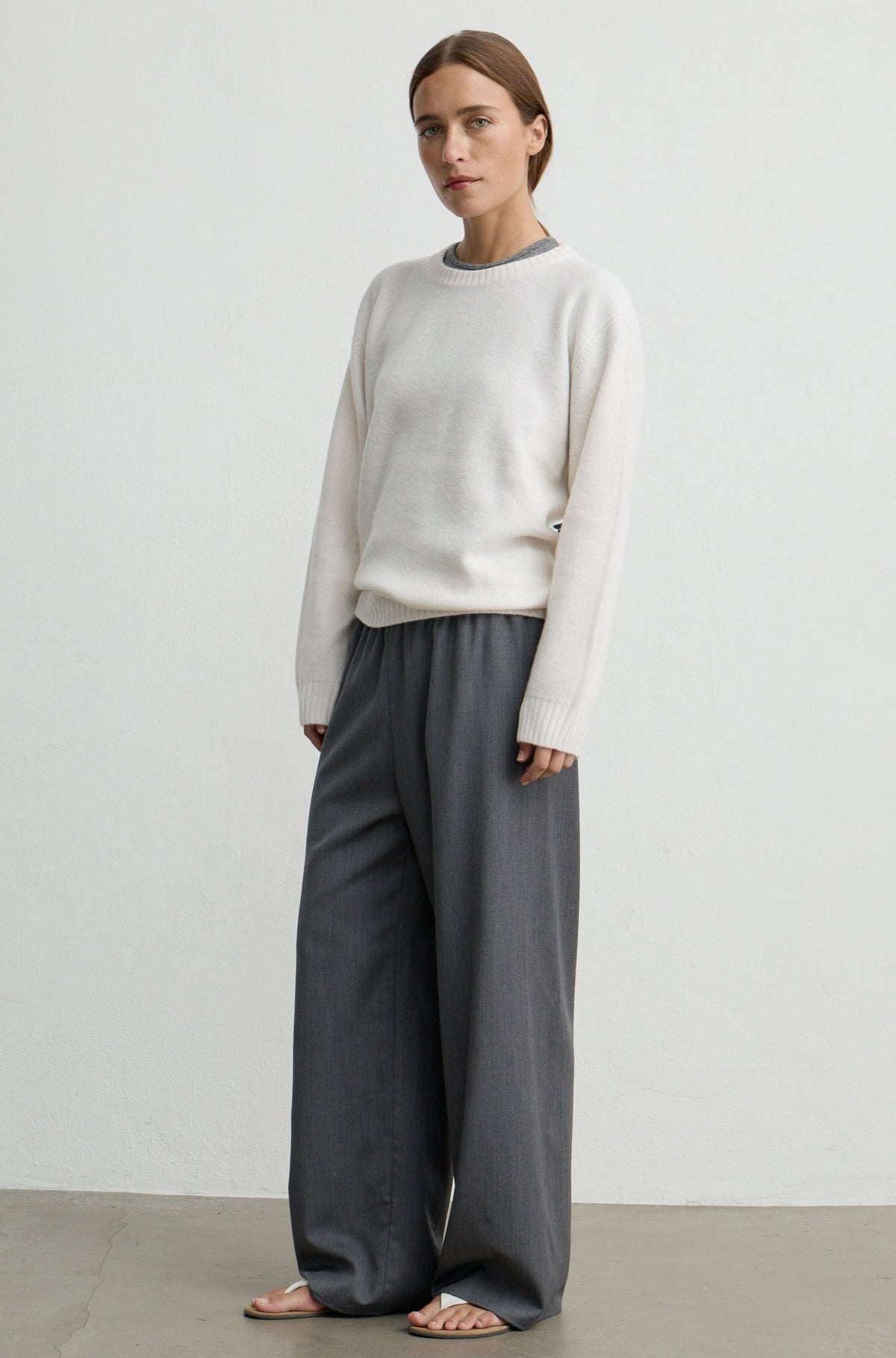 A woman modeling a cream knit crewneck sweater and dark gray wide-leg trousers, standing against a minimalist off-white studio wall.