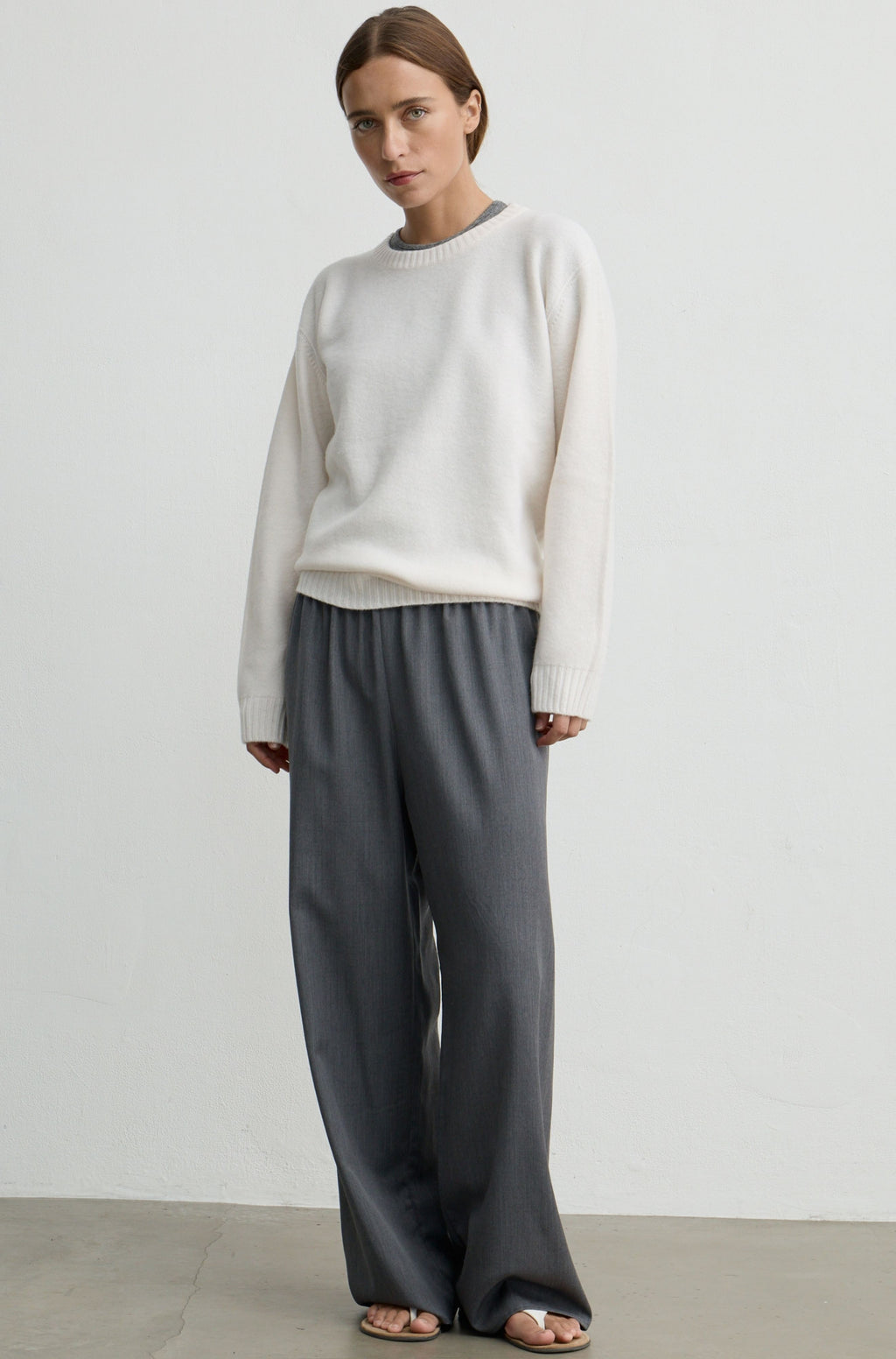 A woman modeling a cream knit sweater and dark gray wide-leg trousers against a plain light gray studio background.