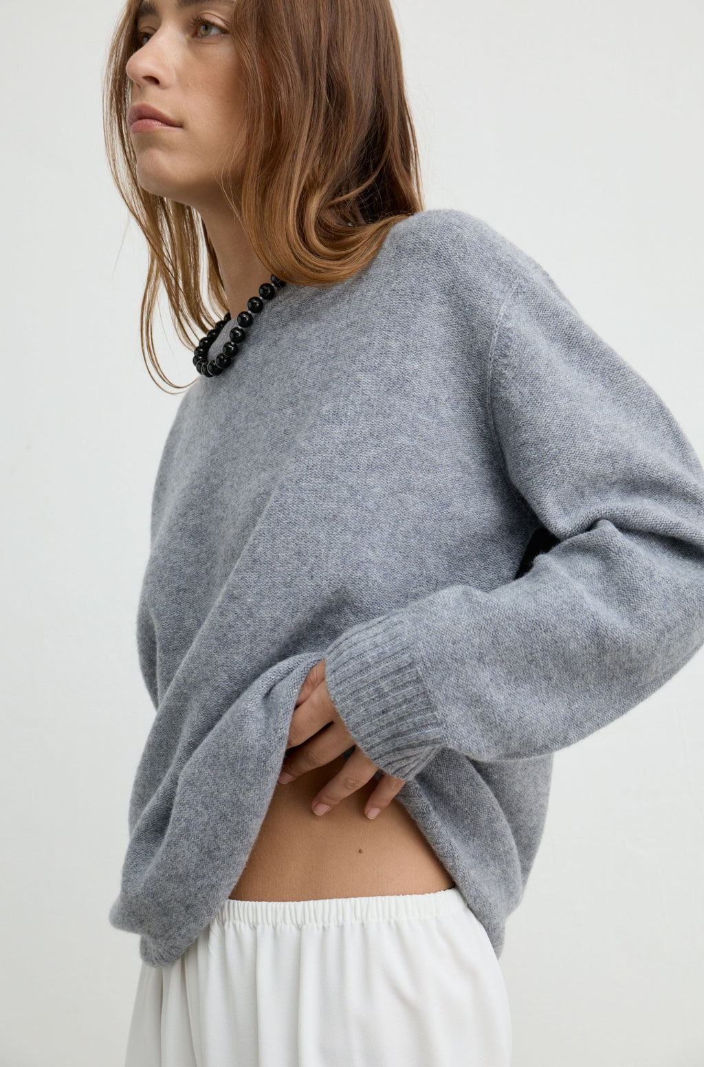 A woman wearing a light gray knit sweater and white pants, shown from the side against a plain light background.