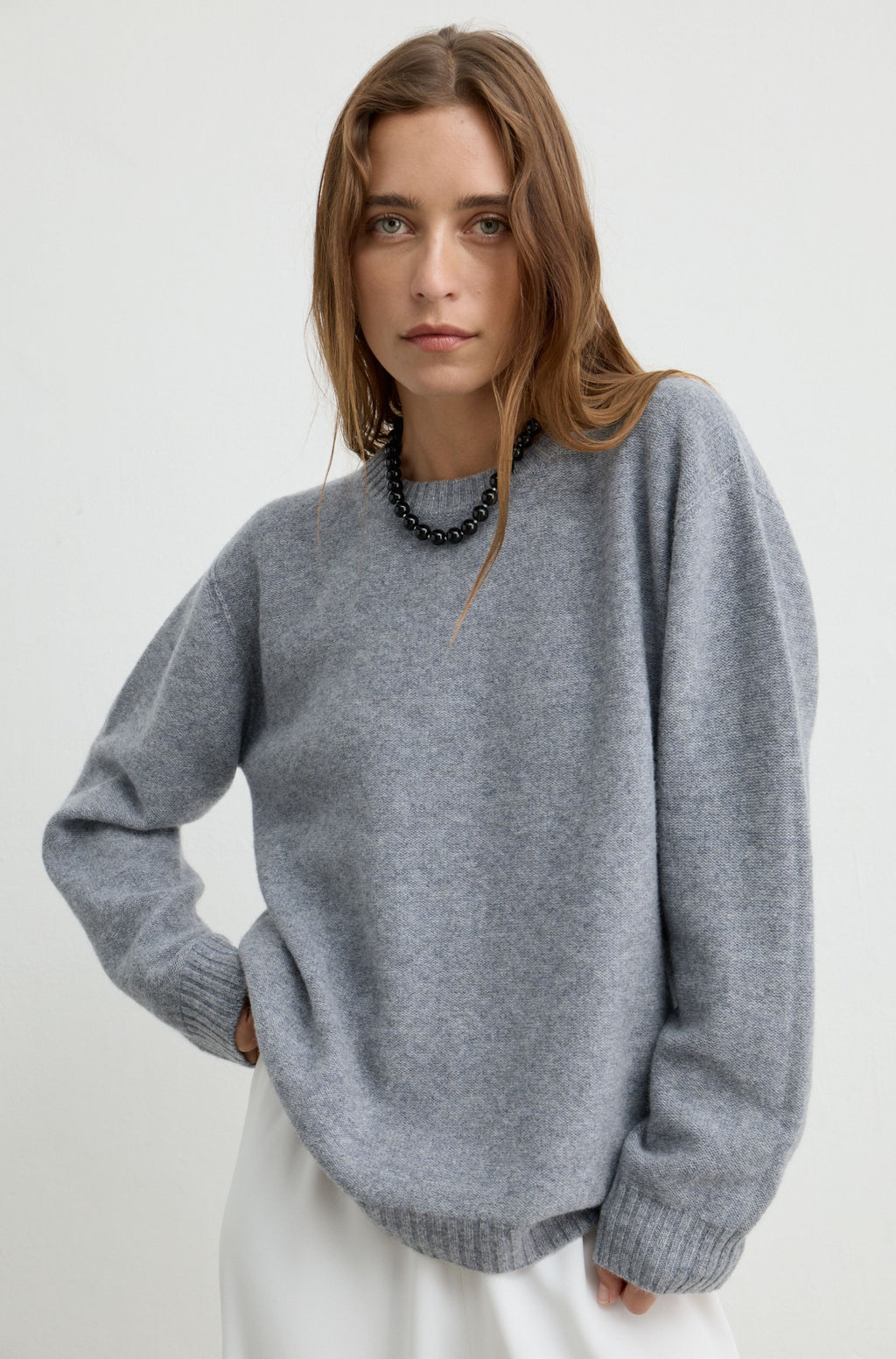A woman wearing an oversized light gray knit sweater with a crew neck and ribbed cuffs, paired with a black beaded necklace and white bottoms, photographed against a plain light gray studio background.