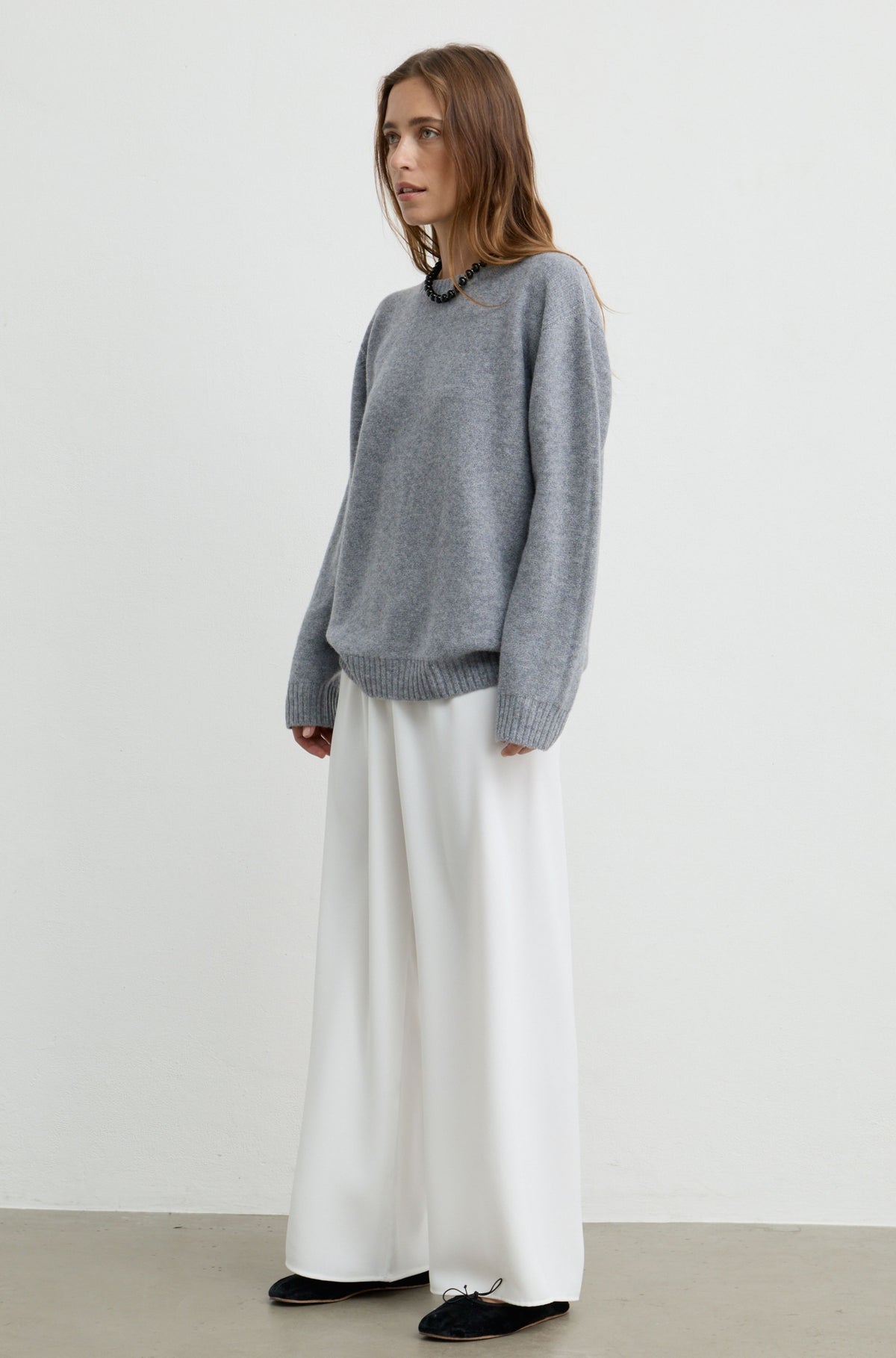 A woman modeling an oversized light gray knit sweater paired with wide-leg white trousers and black shoes, standing against a plain off-white studio wall.
