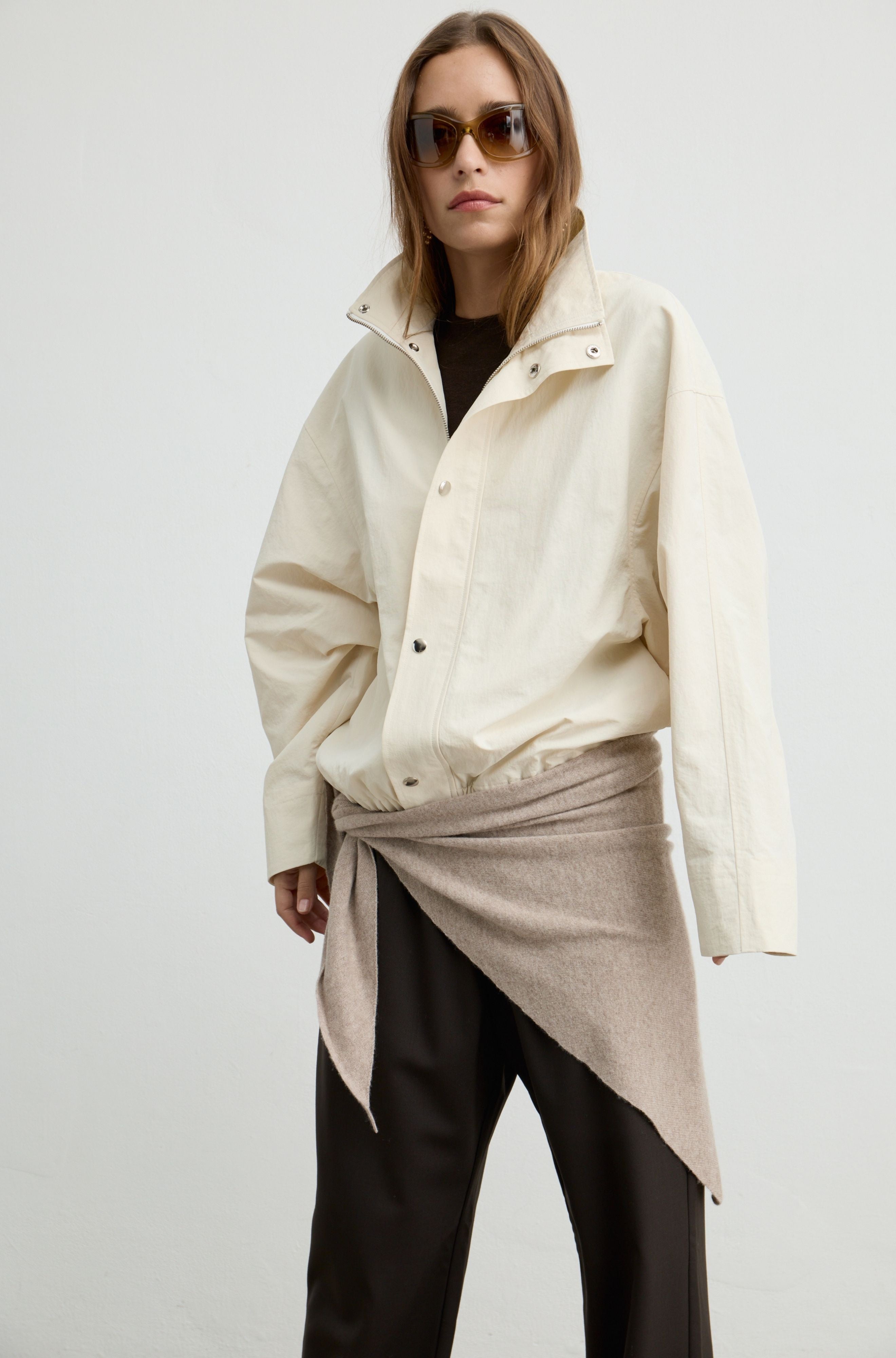 A model wearing an oversized cream-colored jacket with a high collar, styled with a beige wrap shirt and black pants against a plain light gray background.