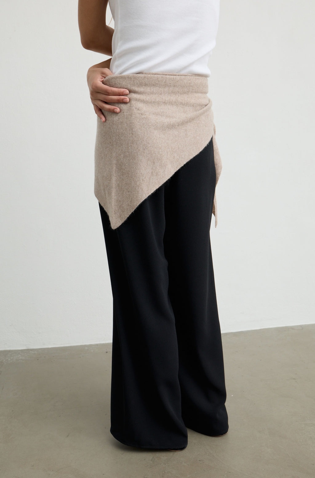 A person shown from the waist down wearing wide-leg black pants with a beige asymmetrical wrap or skirt detail at the waist, paired with a white top, against a plain light gray wall and concrete floor.