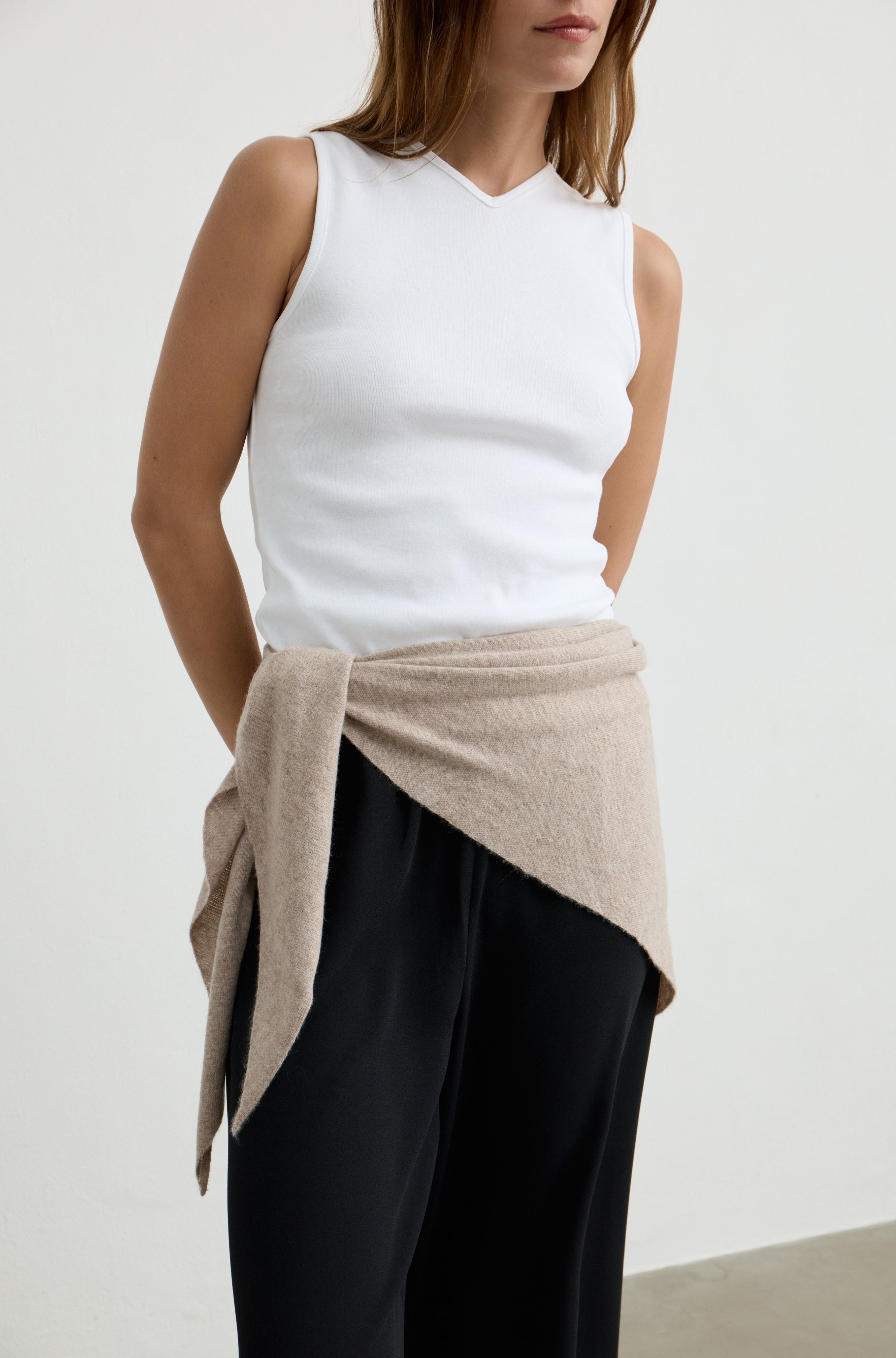 A model wearing a white sleeveless V-neck top, black wide-leg pants, and a beige knit wrap tied around the waist, photographed against a plain light gray studio background.