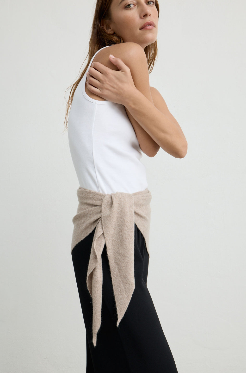 A woman wearing a white tank top and black pants, with a light beige knit wrap tied around her waist, photographed against a plain light gray studio background.