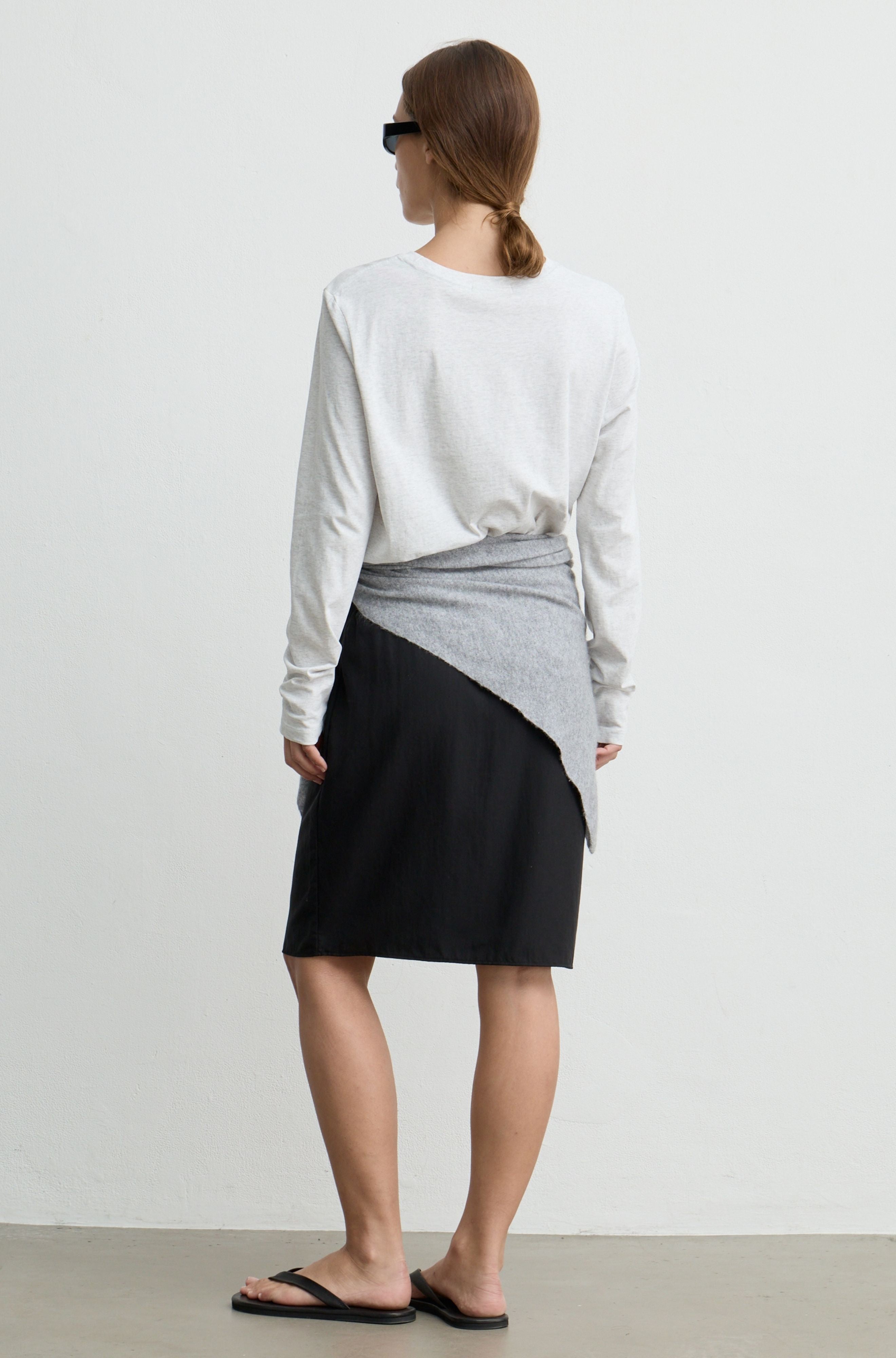 A woman is shown from behind wearing a white long-sleeve top, a black skirt with a gray wrap detail, and black sandals against a plain light gray wall.
