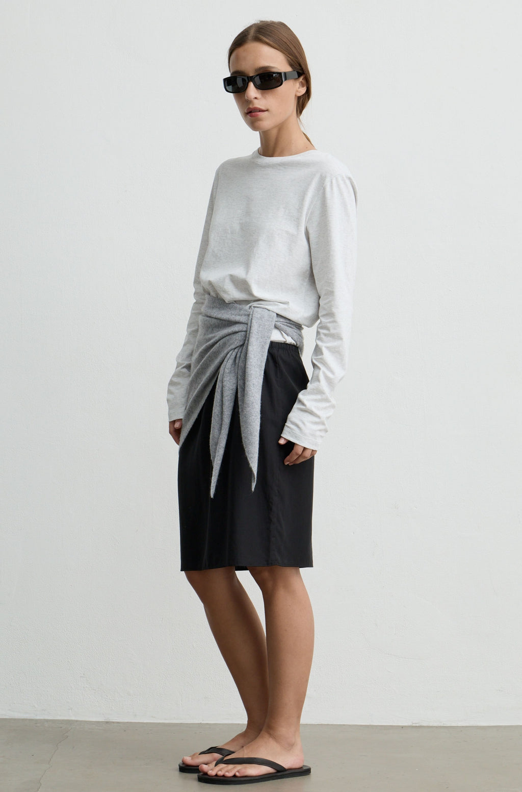 A model stands in a studio wearing a light gray long-sleeve top, a black skirt, and black sandals. A light gray knit wrap is tied around the waist and drapes over the skirt.