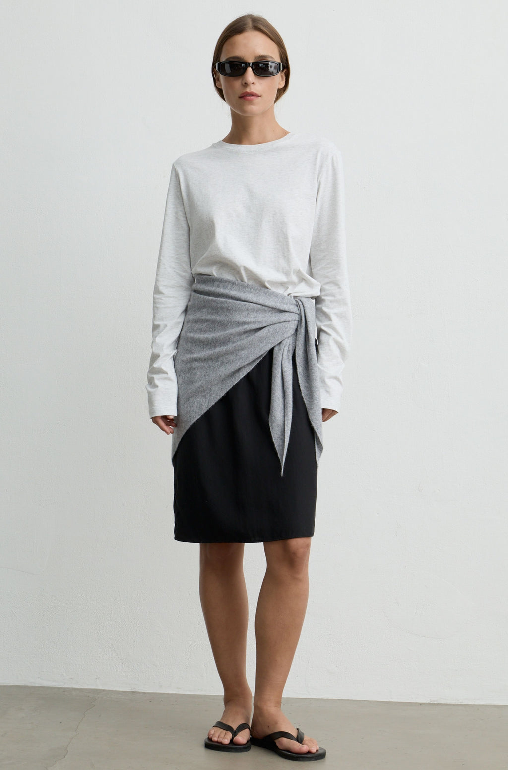 A model stands against a plain light gray wall wearing a white long-sleeve top, a black skirt, and a light gray wrap tied at the waist, with black sandals and sunglasses.