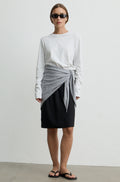 A model stands against a plain light gray wall wearing a white long-sleeve top, a black skirt, and a light gray wrap tied at the waist, with black sandals and sunglasses.
