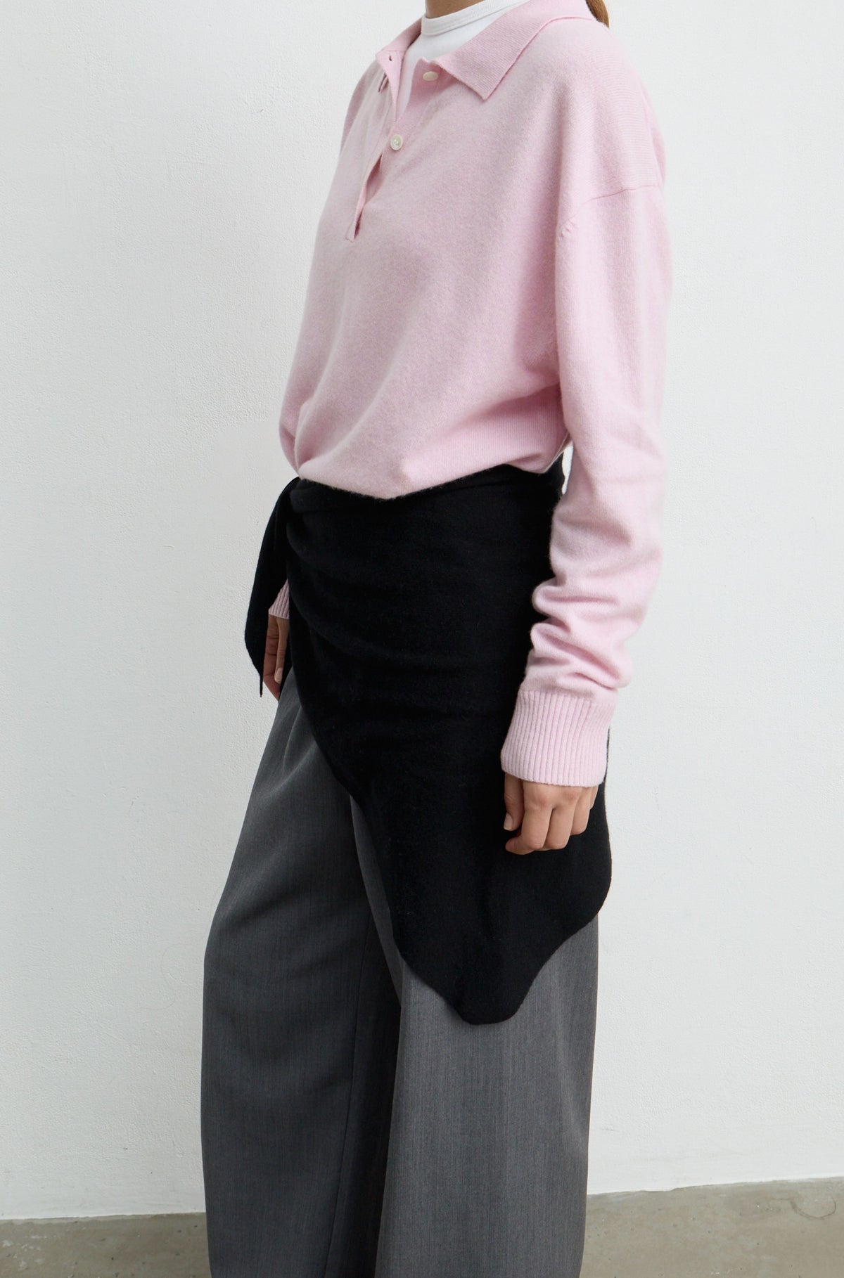 A model wearing a light pink long-sleeve knit polo shirt, a black wrap-style sweater draped over the waist, and wide-leg gray trousers, photographed against a plain off-white wall.