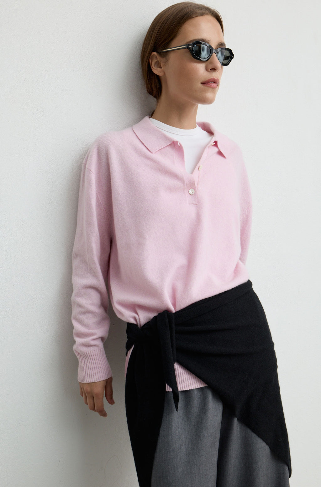 A woman wearing a light pink knit polo sweater over a white undershirt, styled with a black wrap skirt and dark sunglasses, posing against a plain light gray wall.
