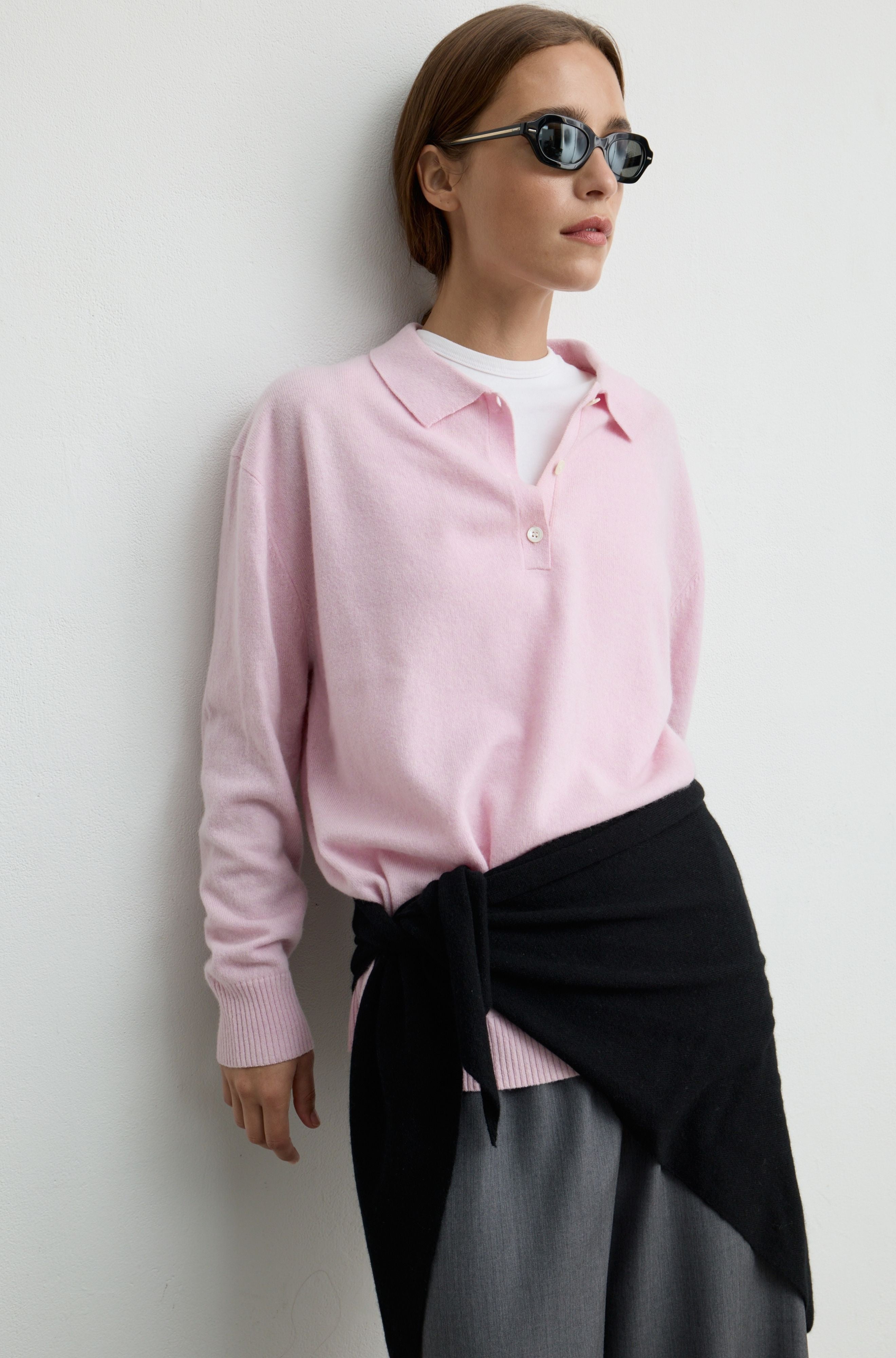 A woman wearing a light pink knit polo sweater over a white undershirt, styled with a black wrap skirt and dark sunglasses, posing against a plain light gray wall.