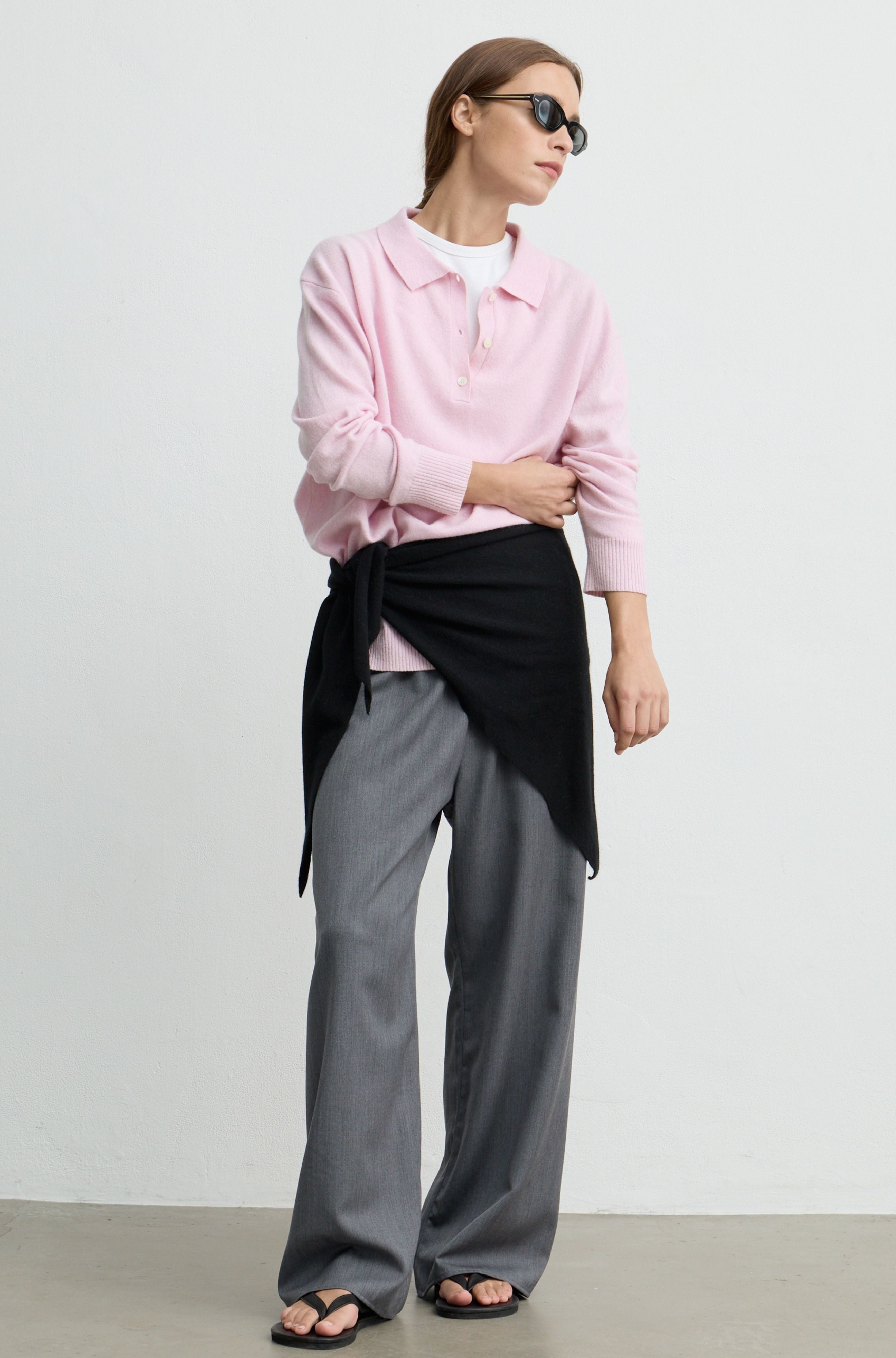 A woman modeling a light pink long-sleeve polo shirt, styled with a black asymmetrical wrap jacket, gray wide-leg trousers, and black sandals against a plain light gray studio background.