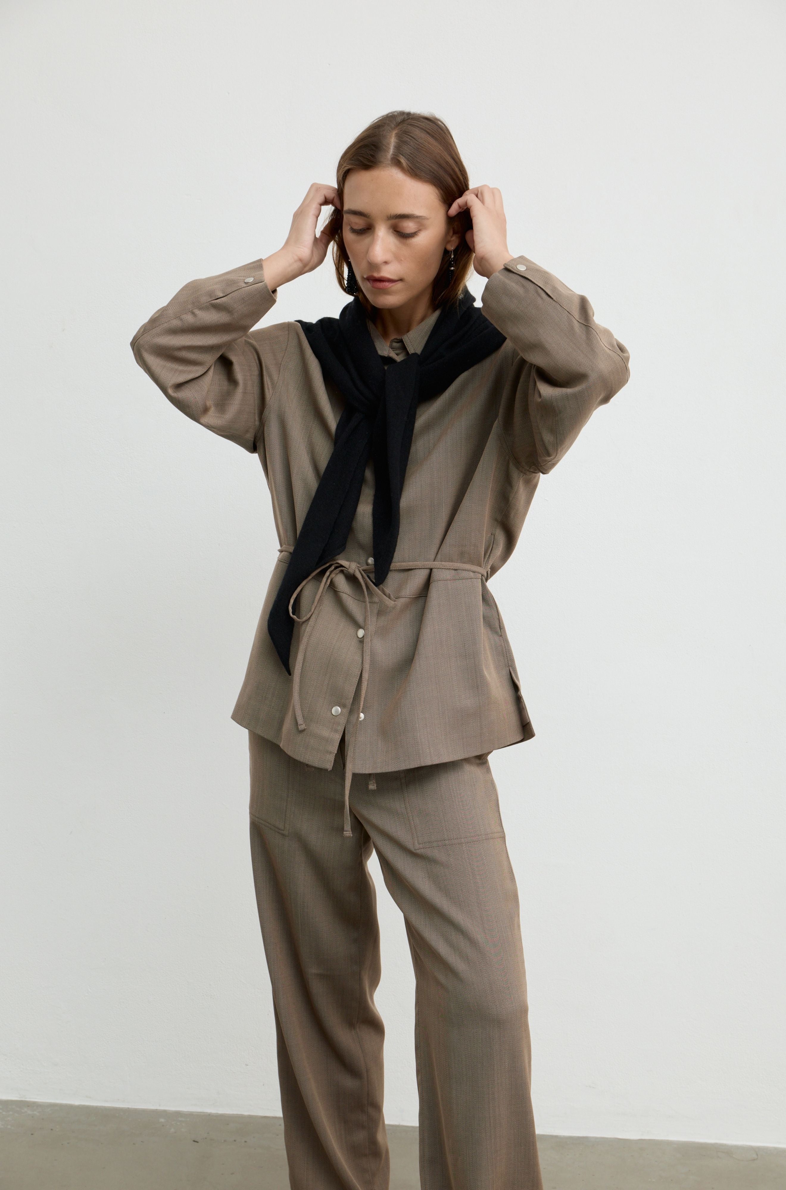 A model wearing a taupe-brown two-piece set consisting of a long-sleeve button-up shirt and matching wide-leg trousers, styled with a black scarf, standing against a plain light gray wall.