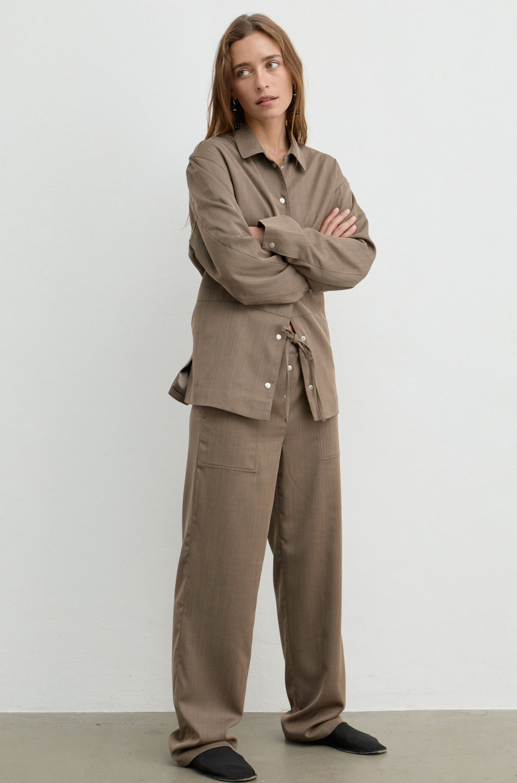 A woman stands with arms crossed wearing a matching taupe-brown two-piece outfit consisting of a long-sleeve button-front shirt with a tie detail at the waist and wide-leg trousers, photographed against a plain light gray studio background.