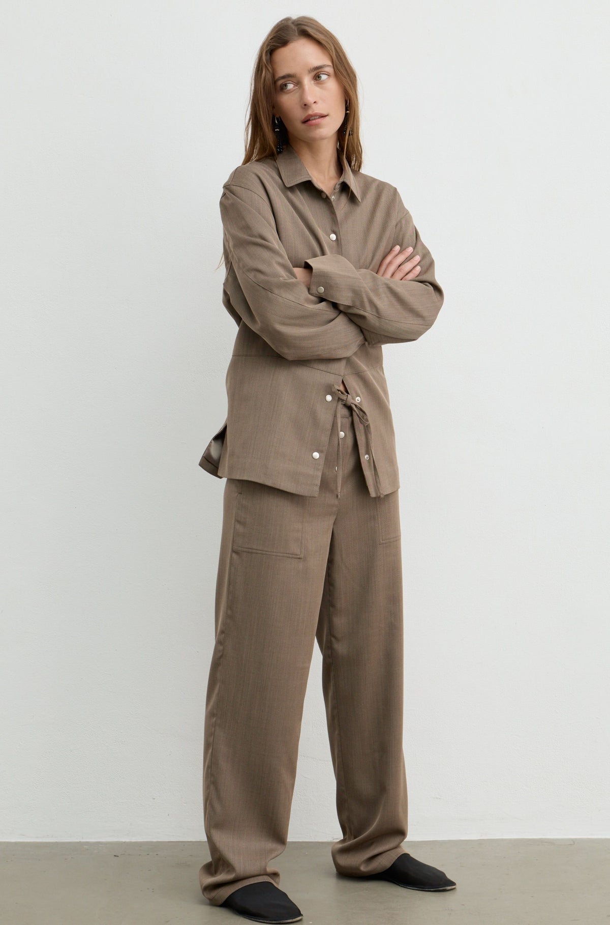 A woman stands with arms crossed wearing a matching taupe-brown two-piece outfit consisting of a long-sleeve button-front shirt with a tie detail at the waist and wide-leg trousers, photographed against a plain light gray studio background.