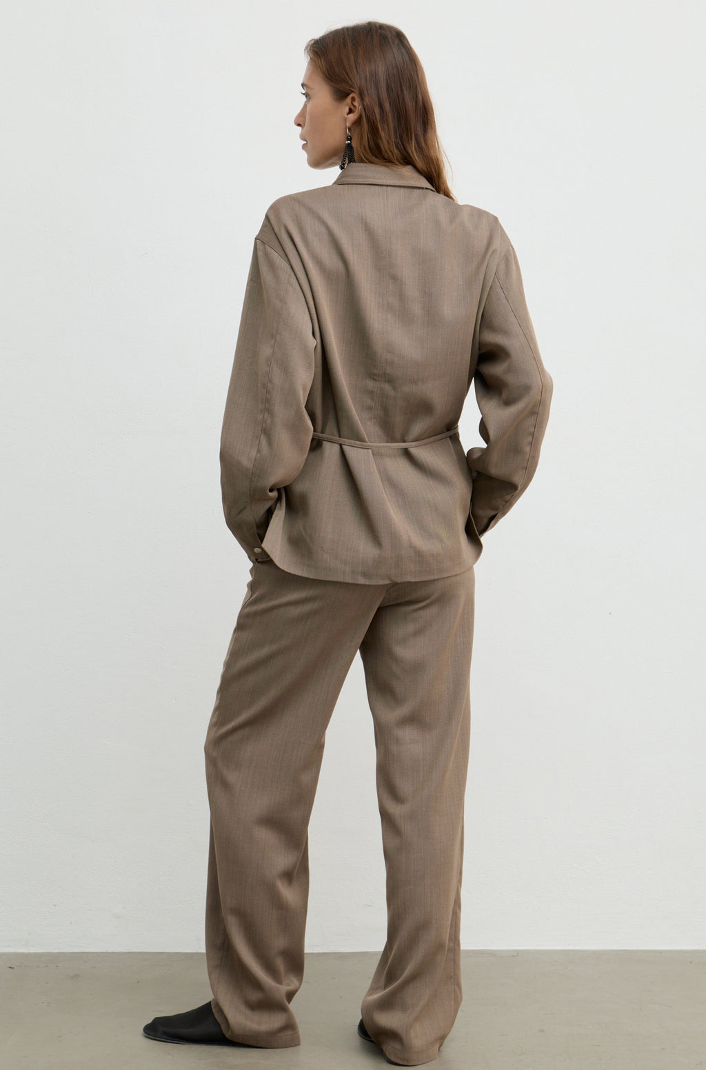 A woman is shown from the back wearing a matching taupe-brown two-piece set consisting of a long-sleeve collared jacket and wide-leg trousers, standing against a plain light gray studio background.