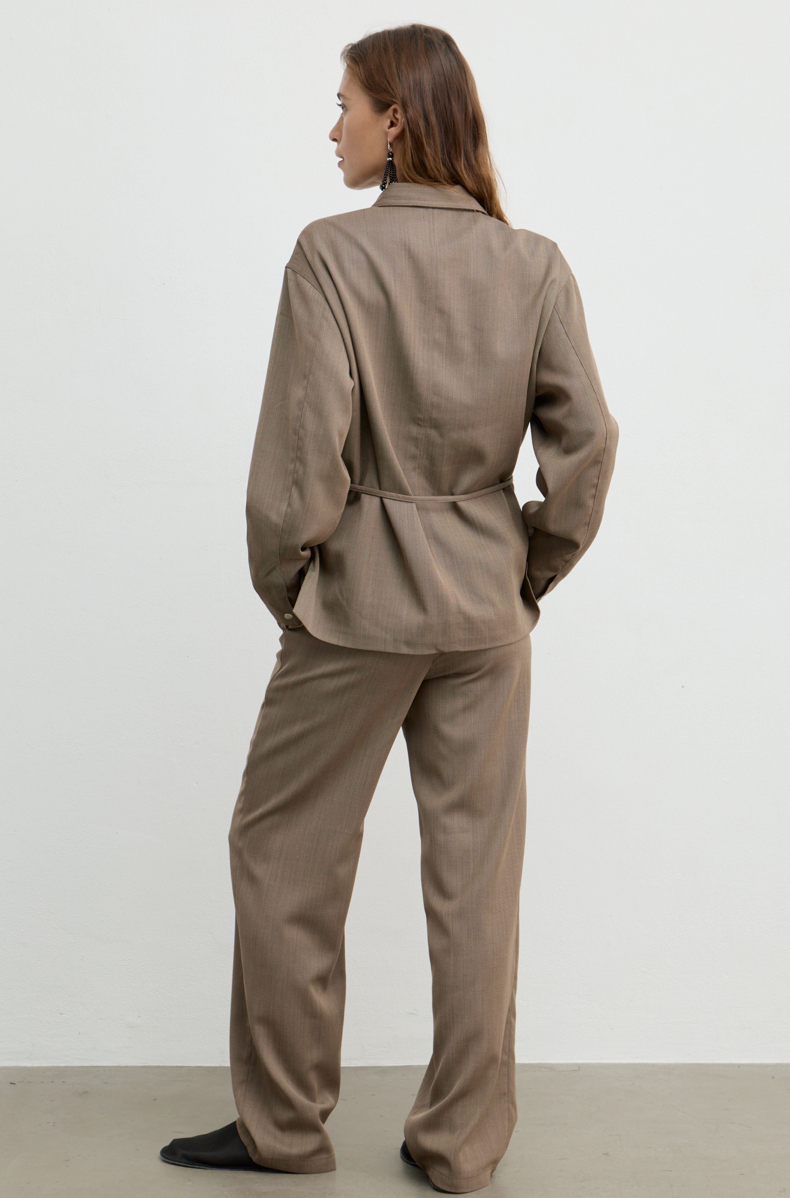 A woman is shown from the back wearing a matching taupe-brown two-piece set consisting of a long-sleeve collared jacket and wide-leg trousers, standing against a plain light gray studio background.