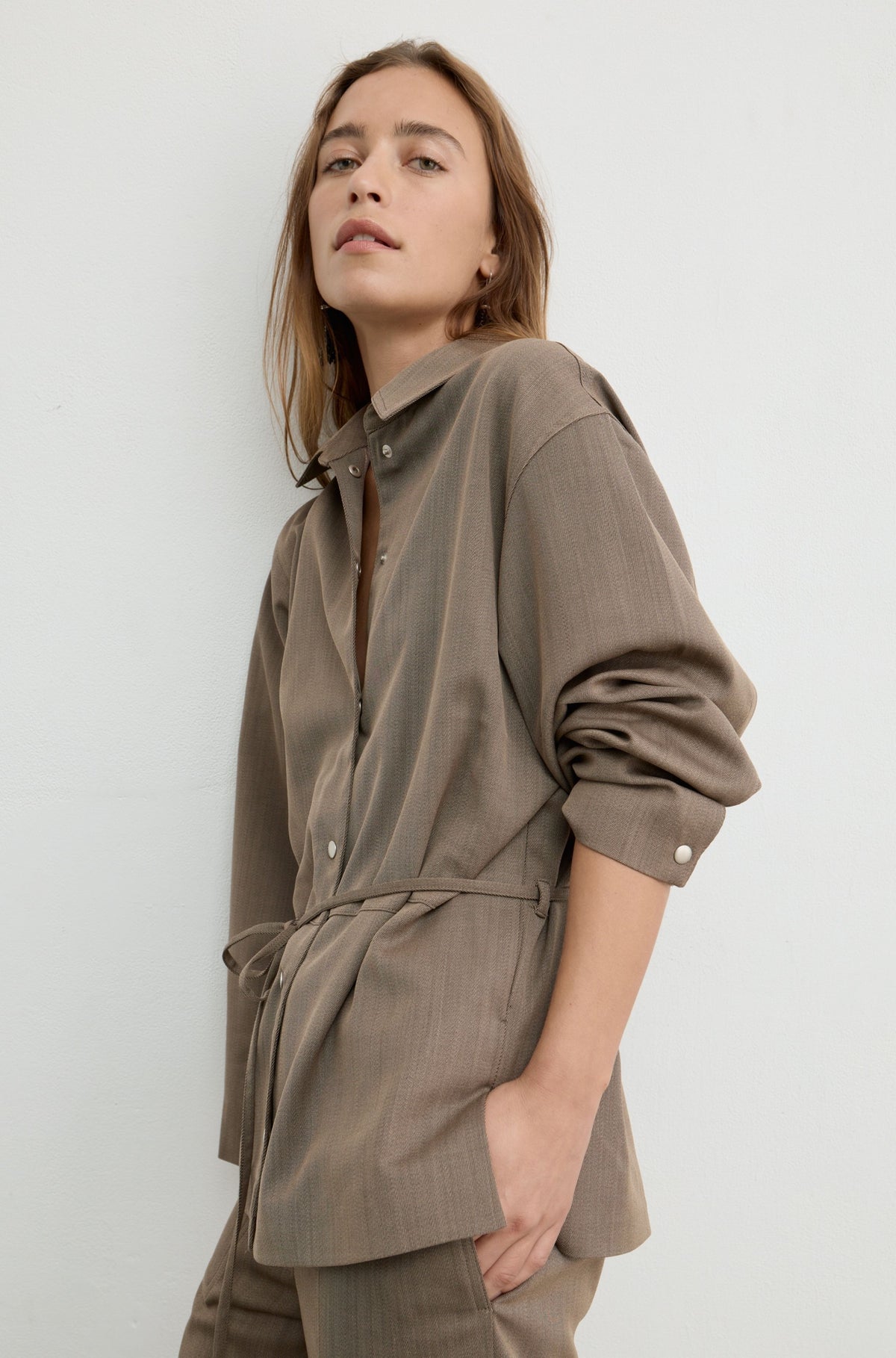 A woman wearing a taupe-brown long-sleeve button-up shirt with a matching tie belt, shown in a studio setting against a light gray background.