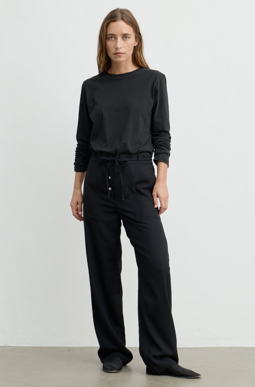 A woman modeling a black long-sleeve crewneck top and matching black wide-leg drawstring pants with button details at the waist, standing against a plain light gray studio background.