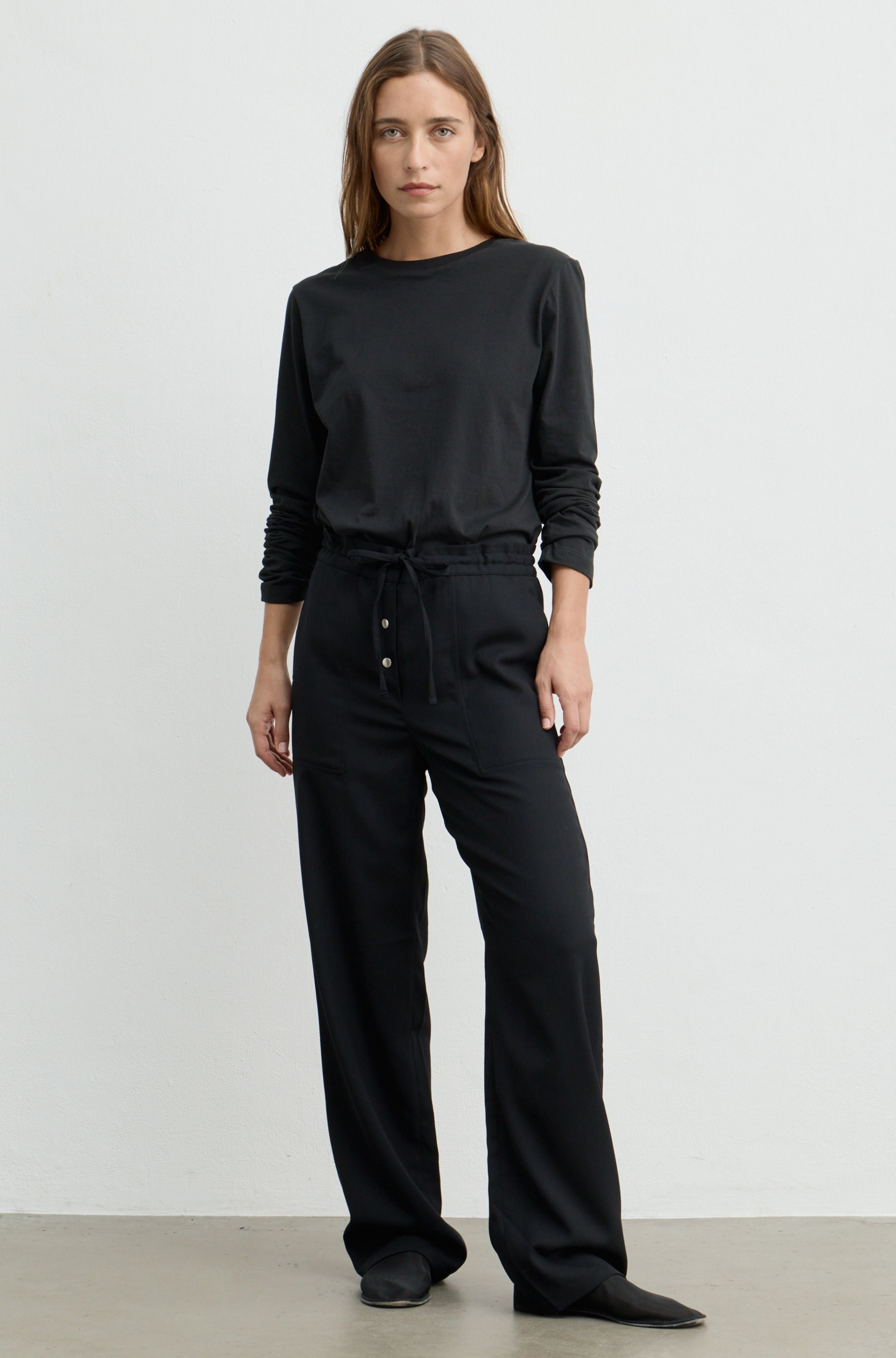 A woman modeling a black long-sleeve crewneck top and matching black wide-leg drawstring pants with button details at the waist, standing against a plain light gray studio background.
