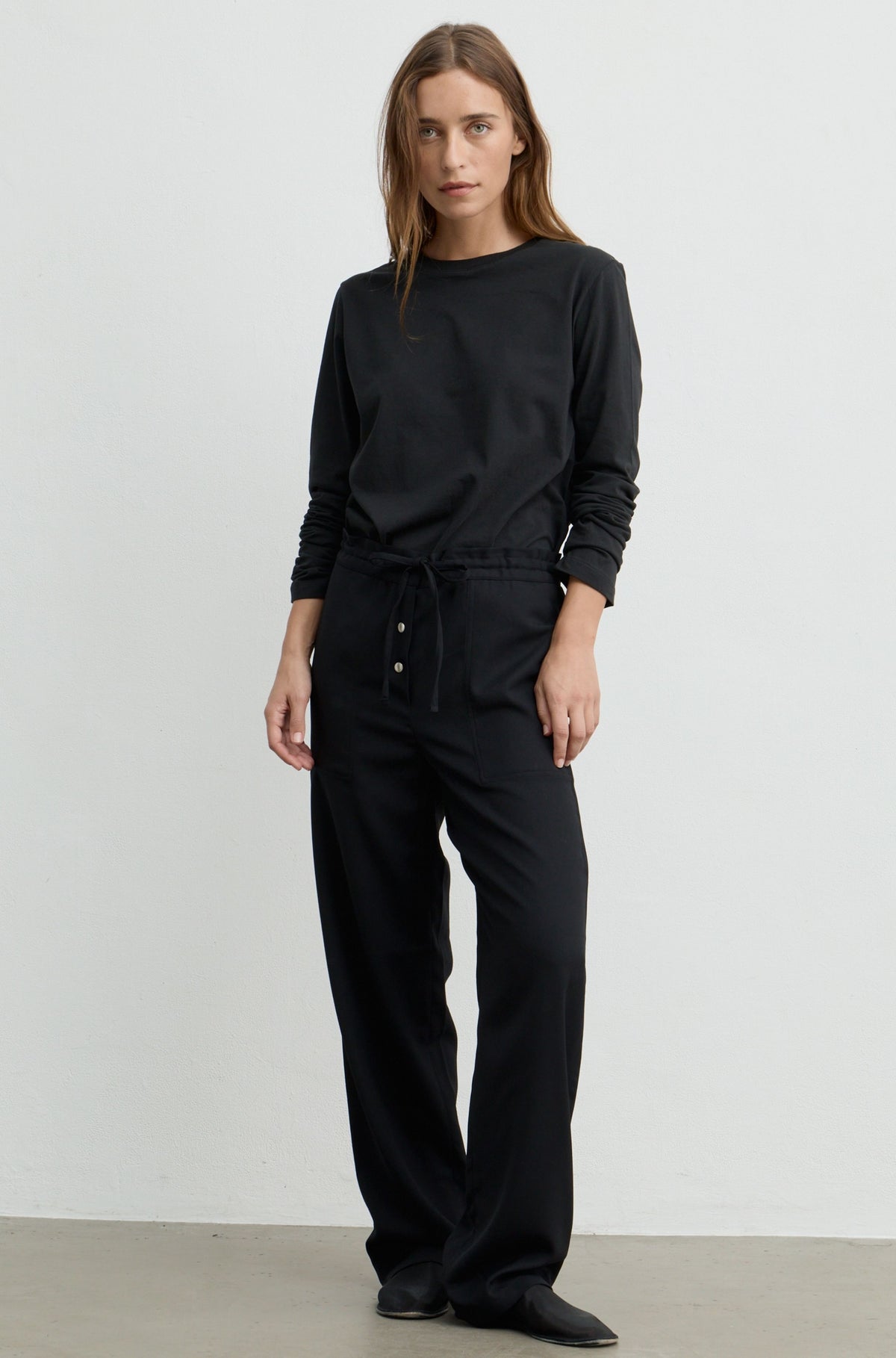 A woman modeling a black long-sleeve crewneck sweatshirt and matching black wide-leg drawstring pants with front buttons, standing against a plain light gray studio wall.