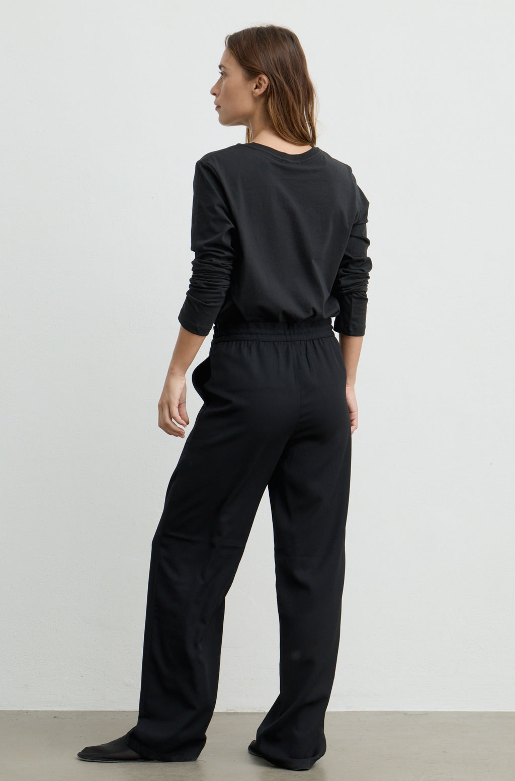 A woman wearing a black long-sleeve top and matching black wide-leg pants, shown from the back against a plain light gray studio background.