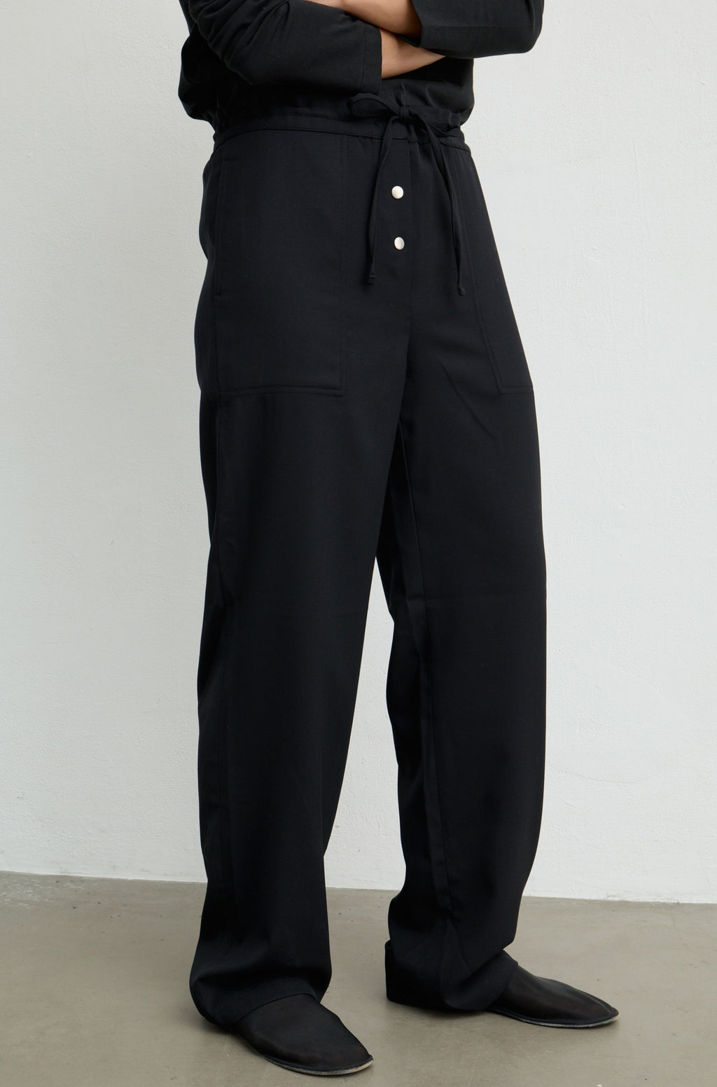 A person wearing loose black drawstring trousers with front buttons, shown from the waist down against a plain light gray wall and concrete floor.