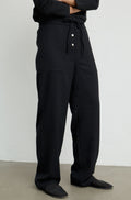 A person wearing loose black drawstring trousers with front buttons, shown from the waist down against a plain light gray wall and concrete floor.