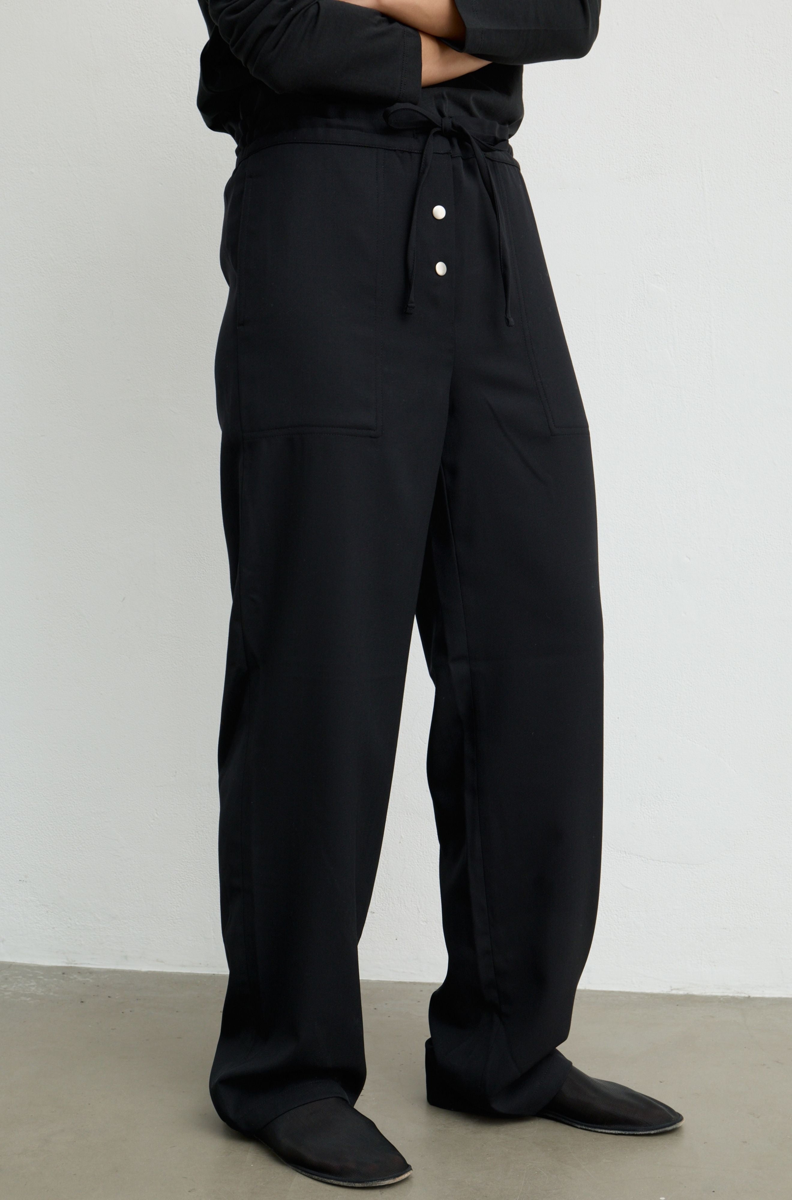 A person wearing loose black drawstring trousers with front buttons, shown from the waist down against a plain light gray wall and concrete floor.