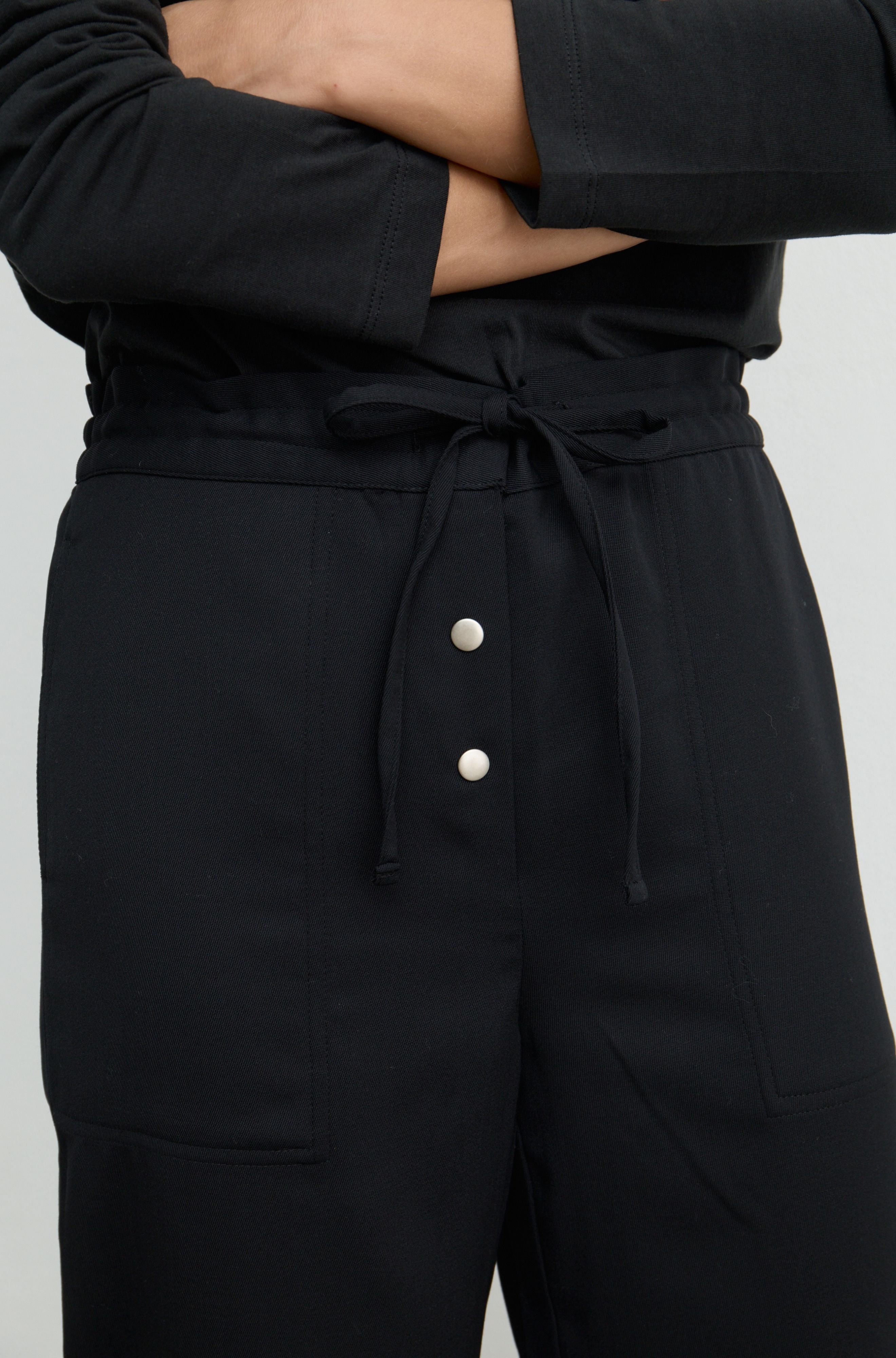 Close-up of a person wearing black wide-leg trousers with a drawstring waist and two silver buttons on the front, paired with a black long-sleeve top against a light gray background.
