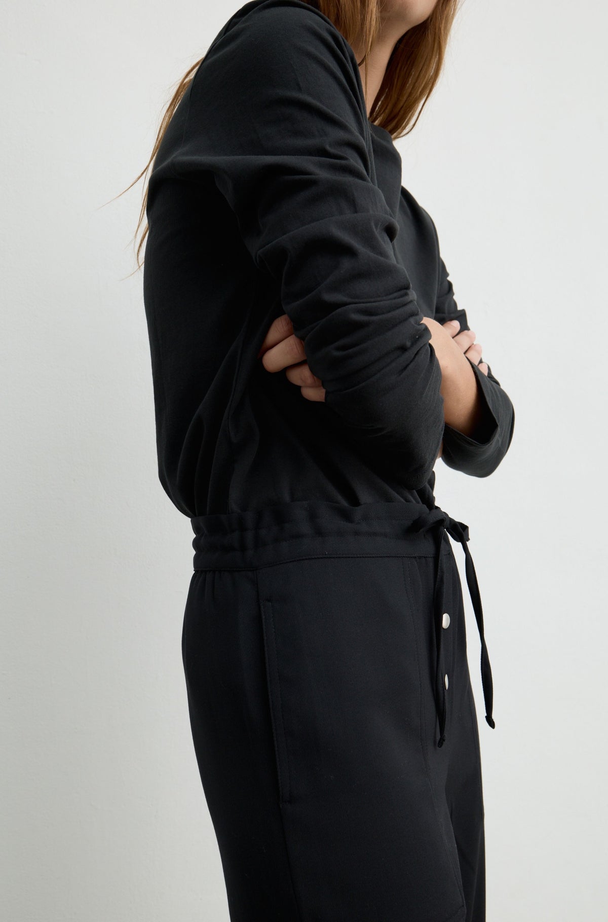 A person wearing a black long-sleeve top and matching black pants with a drawstring waist, shown in a side profile against a light gray studio background.