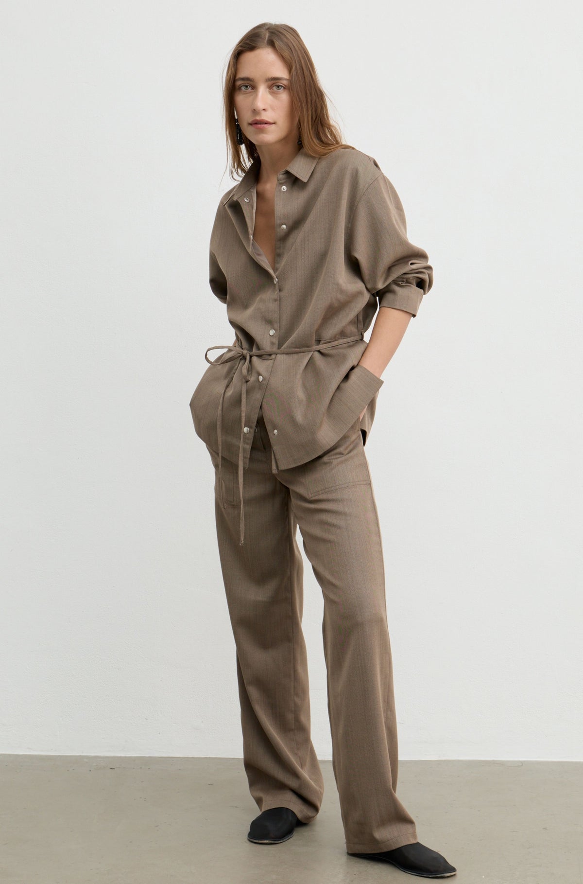 A woman modeling a matching taupe-brown two-piece linen set consisting of a long-sleeve button-down shirt with a tie waist and matching wide-leg trousers, set against a neutral studio background.