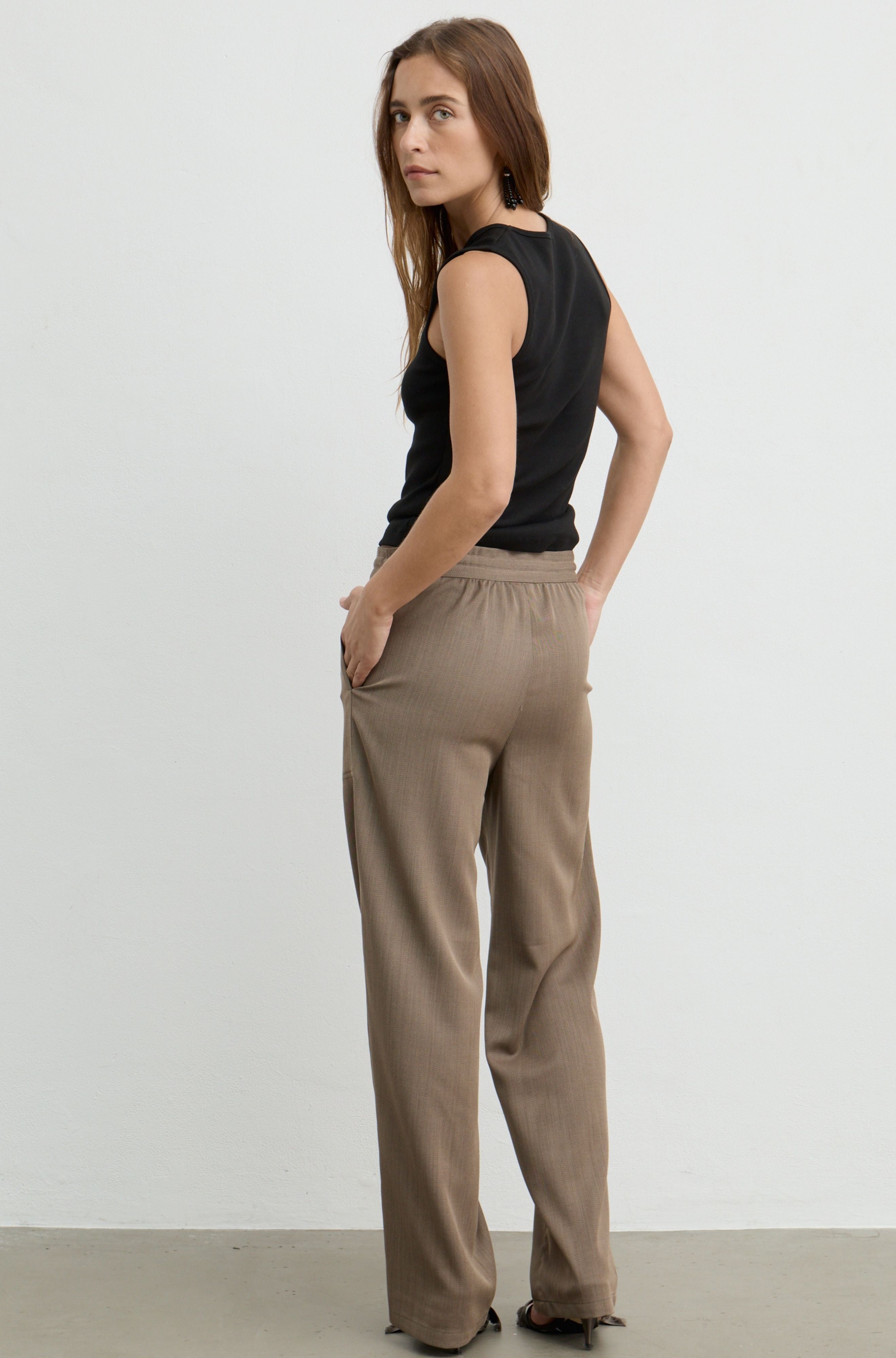 A woman wearing beige wide-leg pleated trousers and a black sleeveless top, photographed from the back against a plain light gray studio wall.