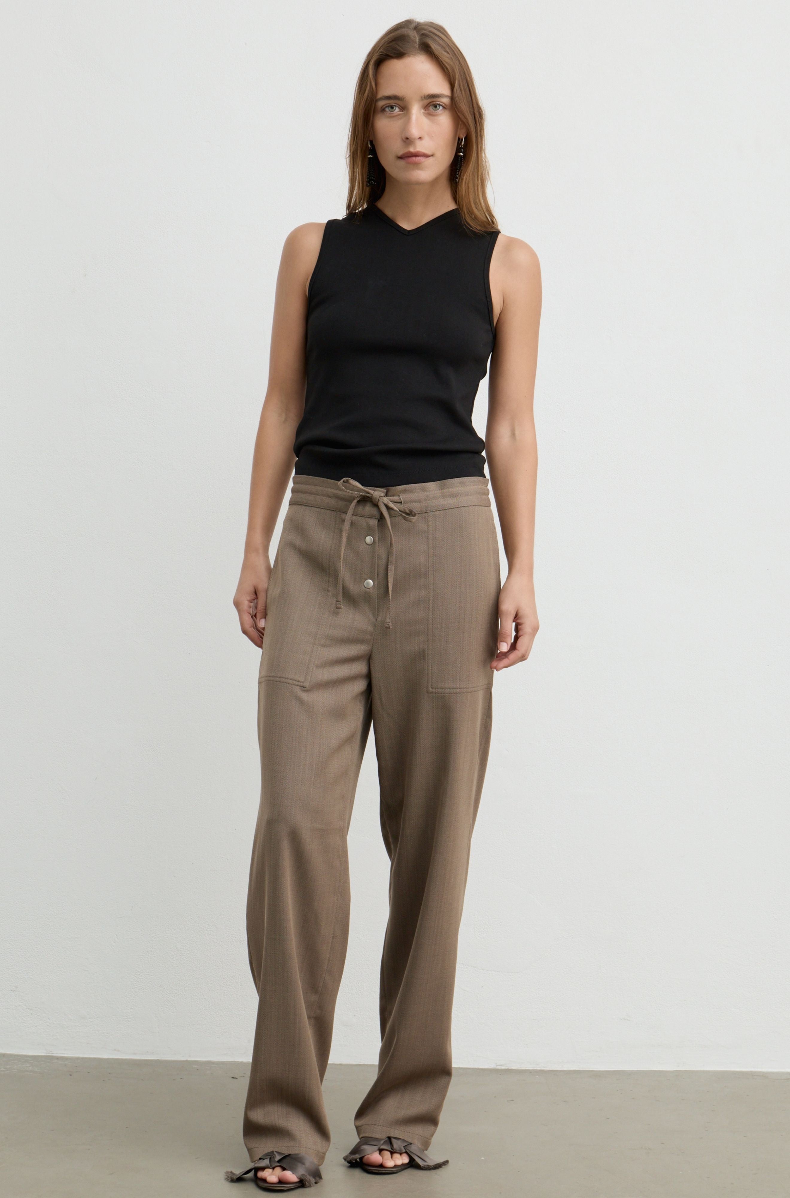 A woman modeling a black sleeveless mock-neck top and brown wide-leg drawstring pants against a plain light gray studio background.