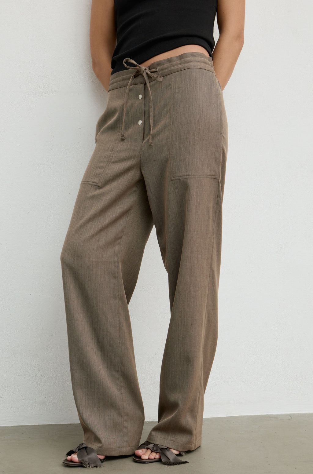 A person wearing loose, wide-leg taupe-brown drawstring pants with side pockets, paired with a black sleeveless top and black sandals, standing against a plain light gray wall.
