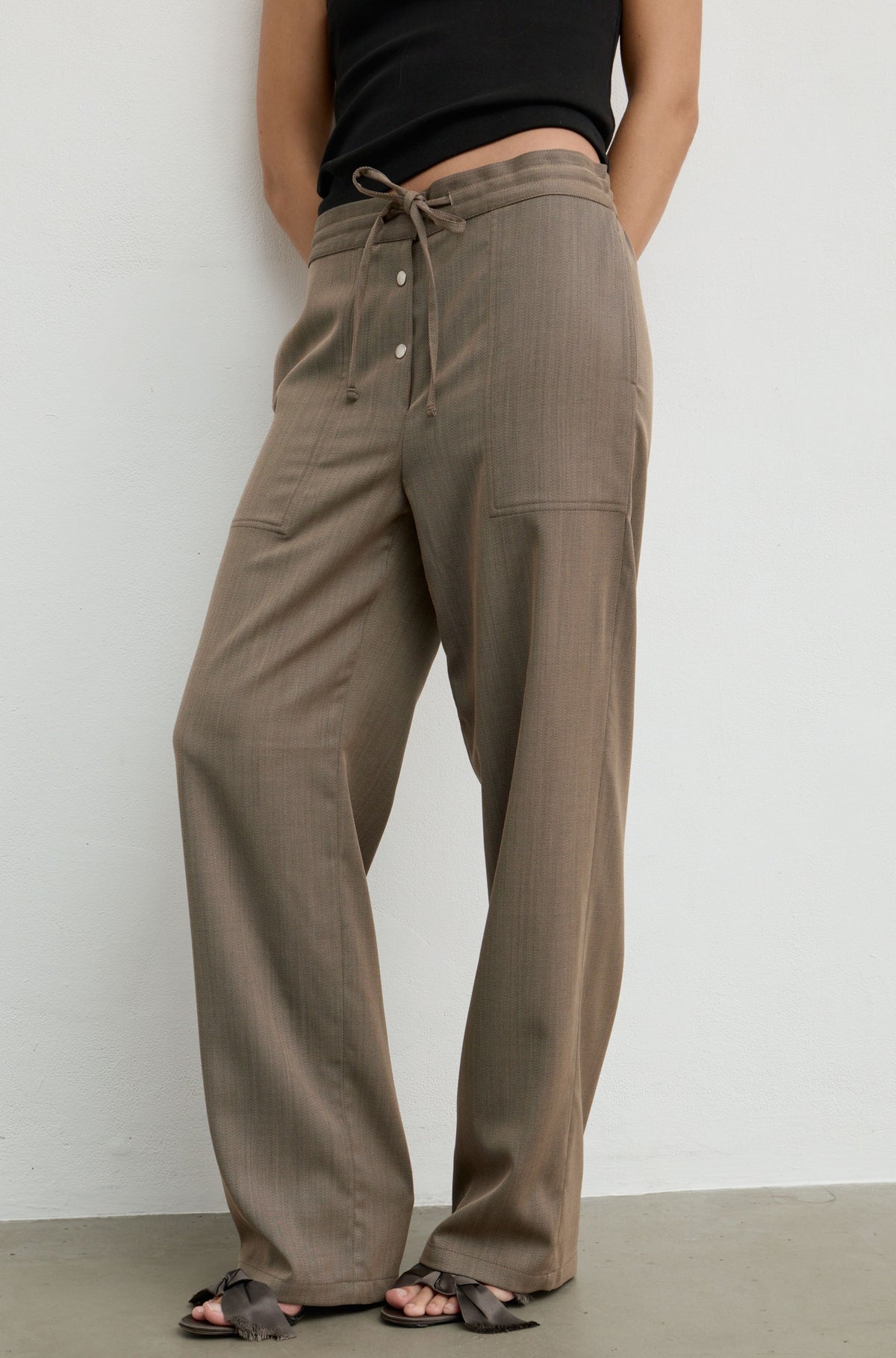 A person wearing loose, wide-leg taupe-brown drawstring pants with side pockets, paired with a black sleeveless top and black sandals, standing against a plain light gray wall.