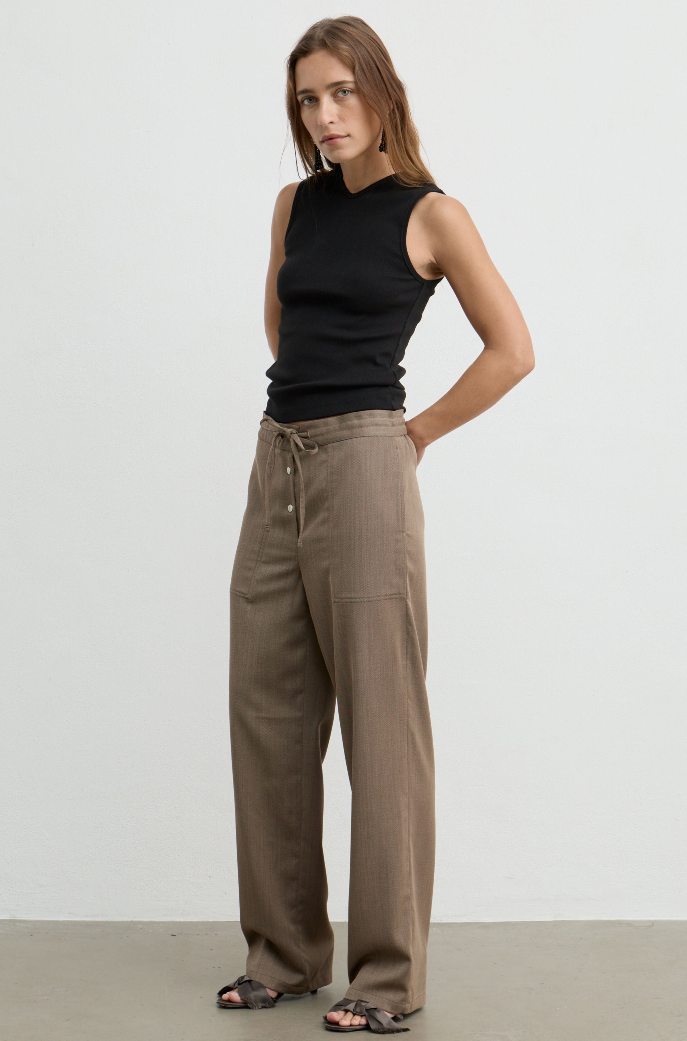 A woman modeling a black sleeveless ribbed tank top and high-waisted, wide-leg taupe-brown pants with a drawstring waist, standing against a plain light gray studio background.