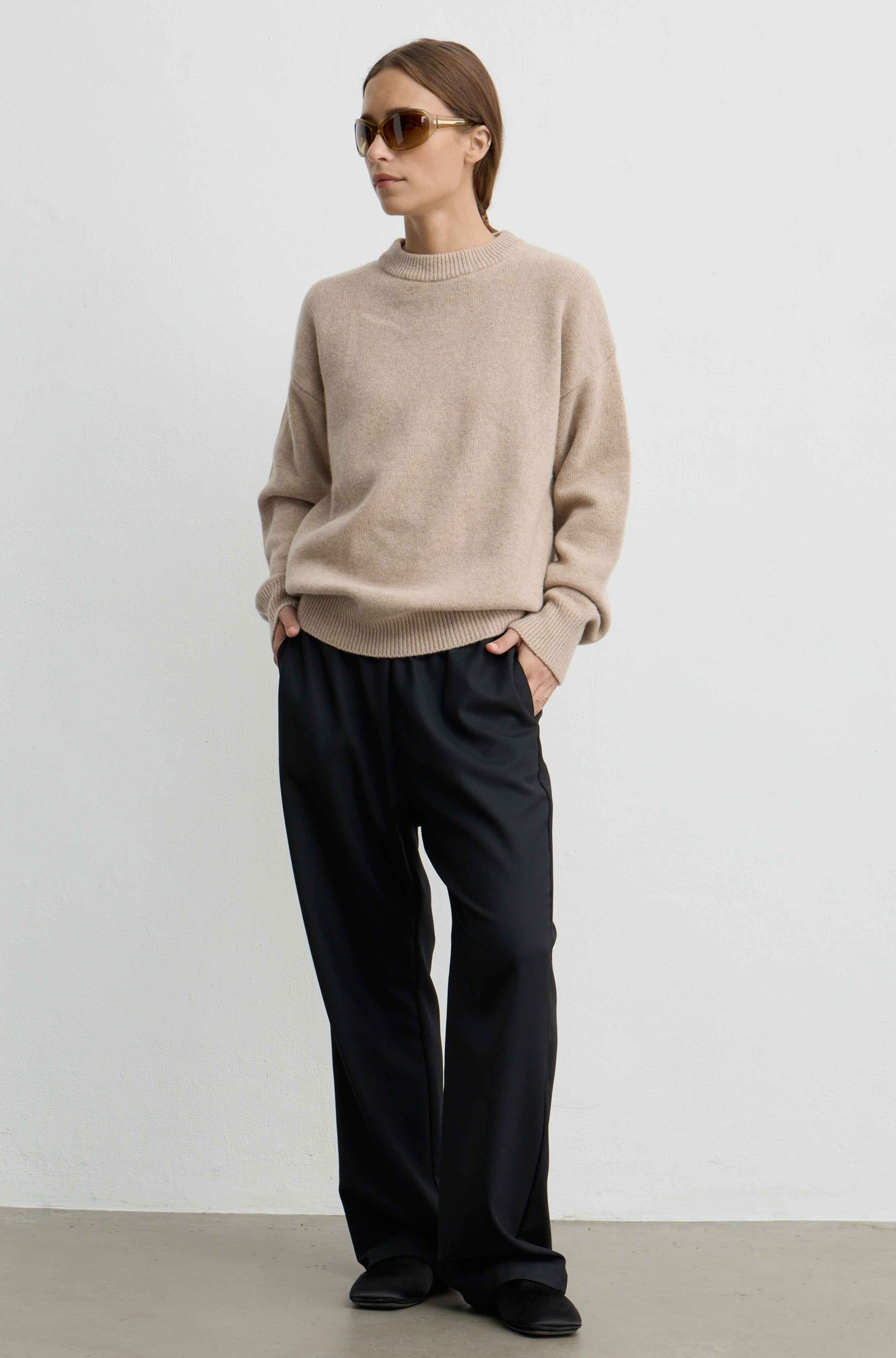 A woman modeling a light beige crewneck knit sweater and black wide-leg trousers, standing against a plain light gray wall.