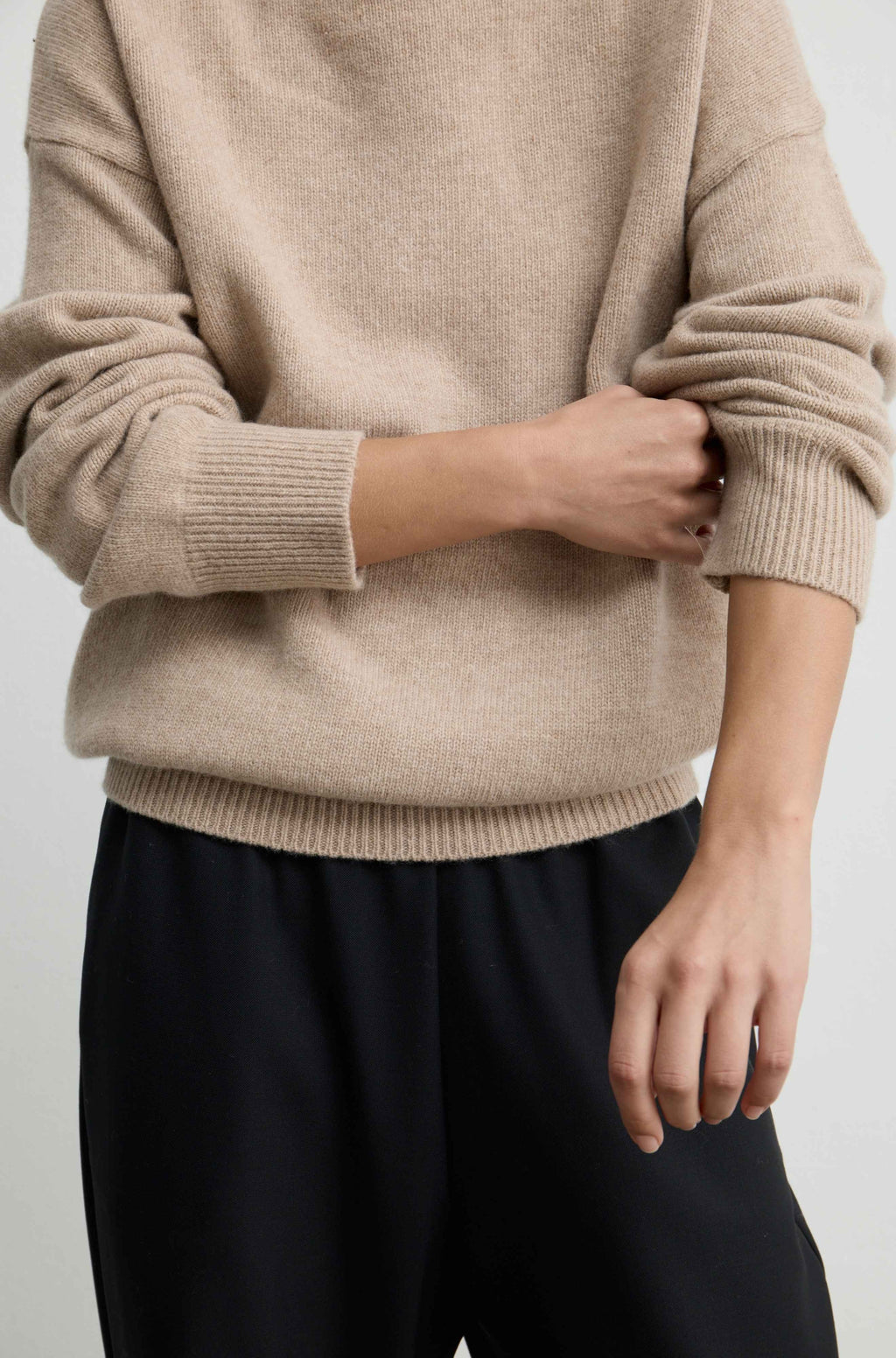 A close-up of a person wearing a beige knit sweater with cuffed sleeves and black pants against a plain light background.
