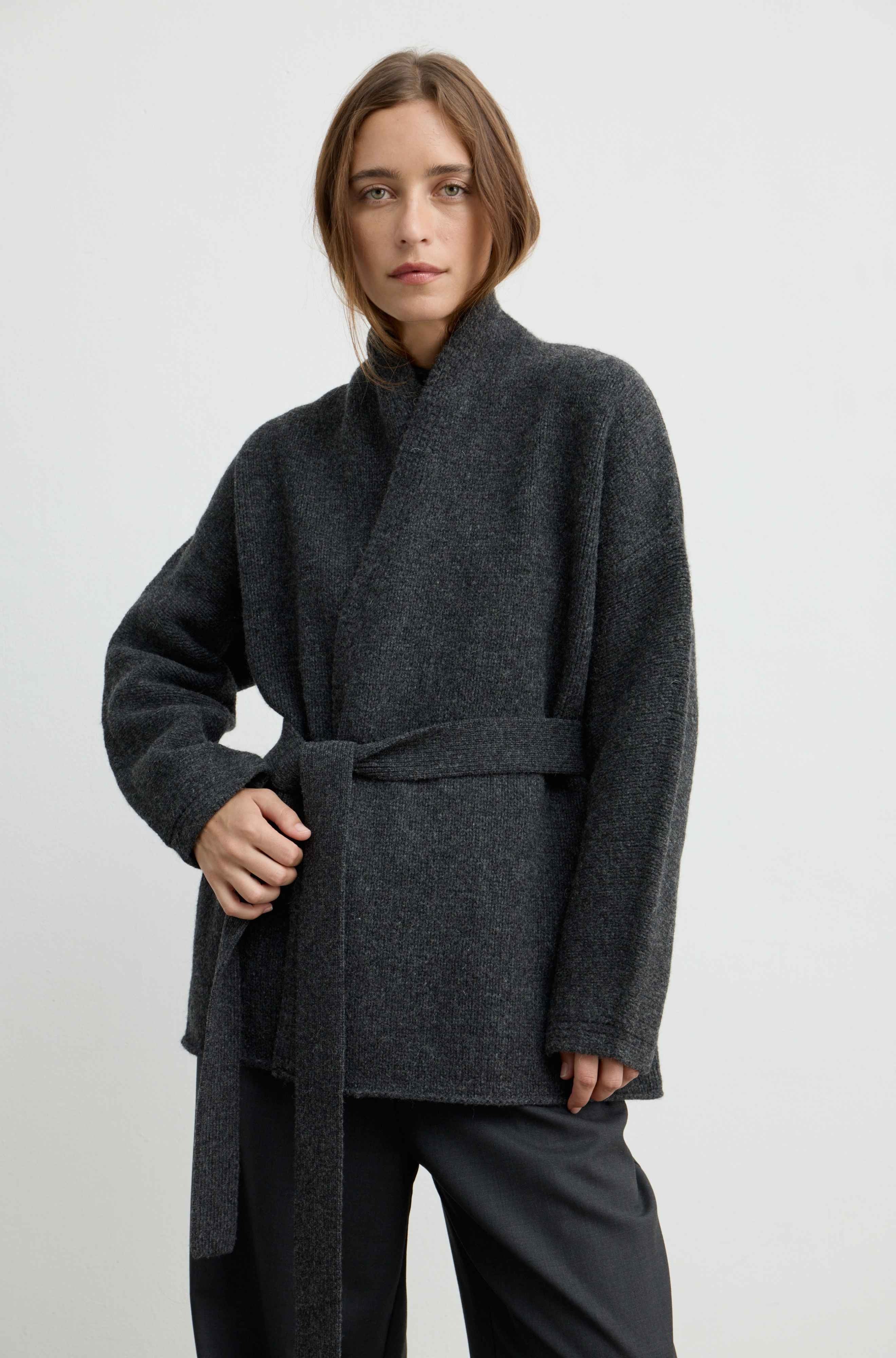 A woman modeling a dark charcoal gray wrap coat with a matching waist tie belt, paired with dark trousers, against a plain light gray studio background.