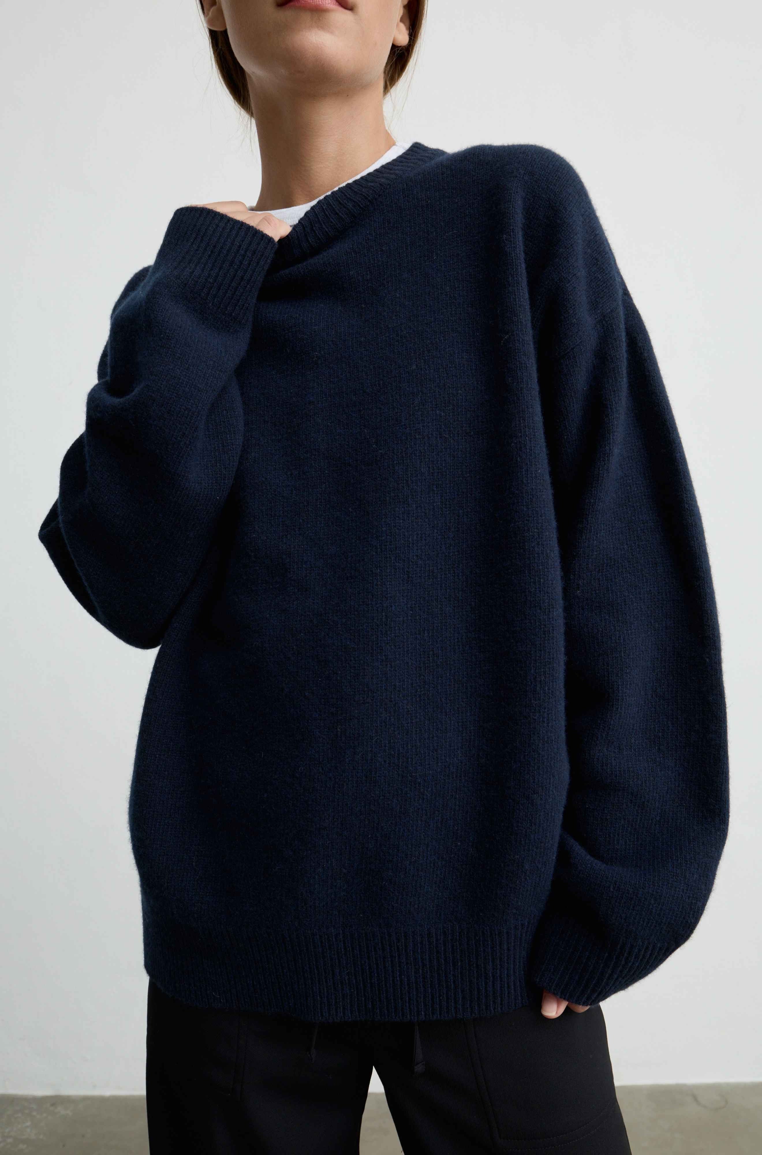 A person wearing an oversized dark navy knit sweater with a wide collar and ribbed cuffs and hem, paired with black pants against a plain light gray wall.