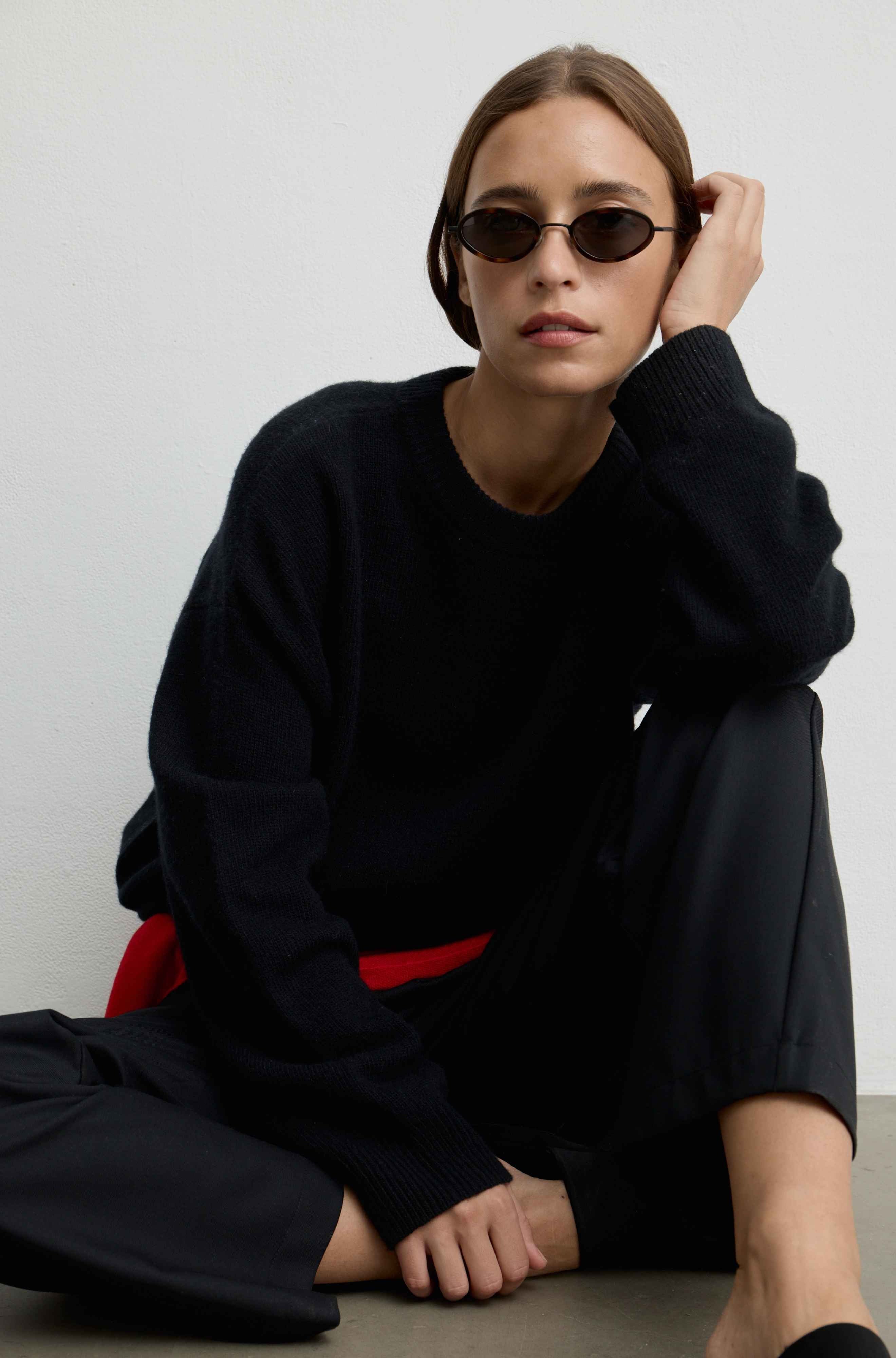 A woman wearing a black knit crewneck sweater, black wide-leg trousers, and black oval sunglasses sits against a plain light gray wall.
