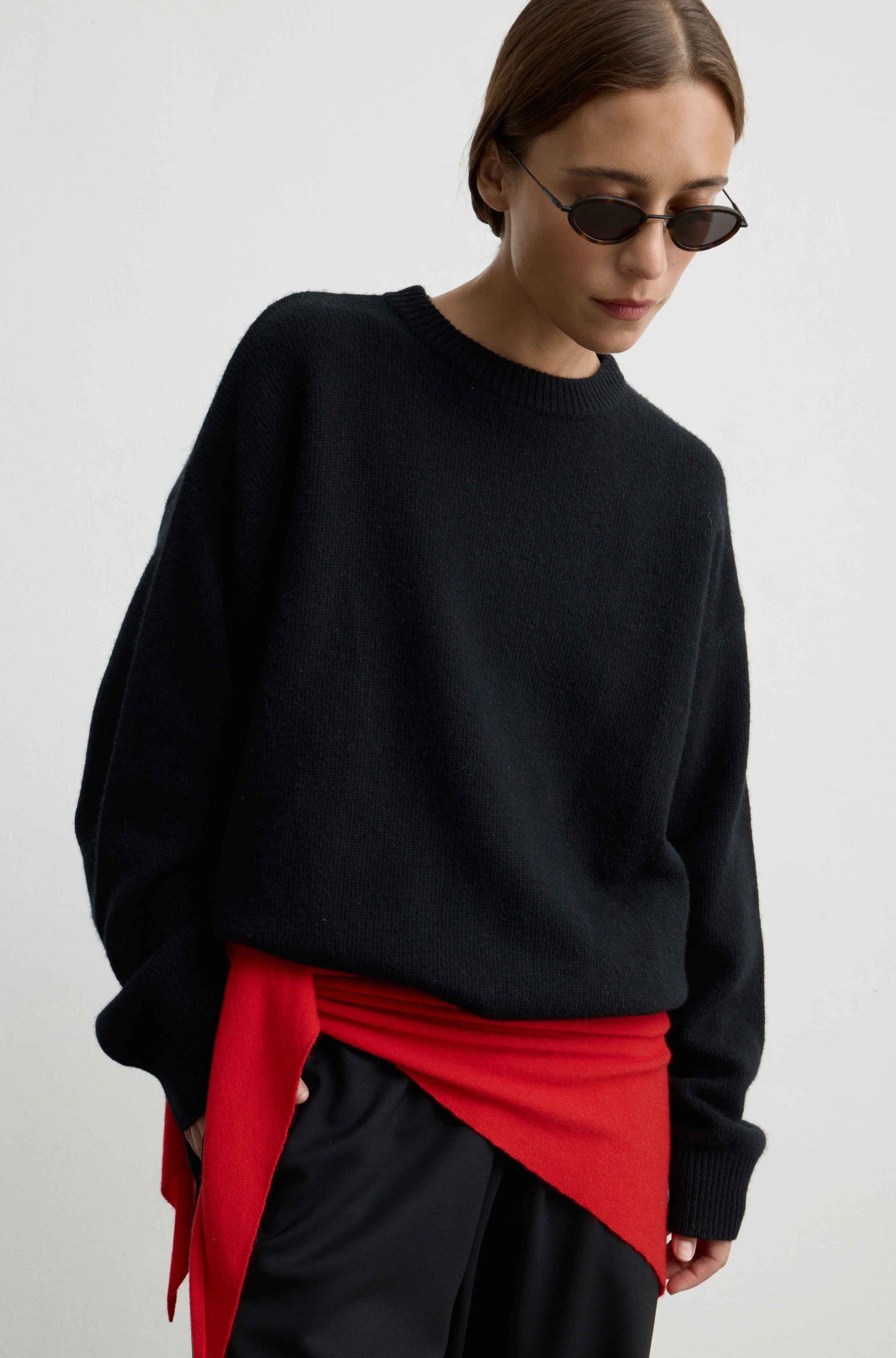A woman wearing an oversized black knit sweater, black trousers, and a bright red wrap tied around her waist, photographed against a plain light background.