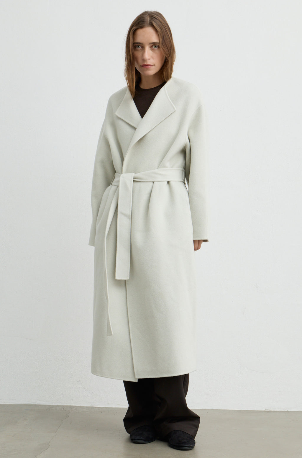 Woman wearing a light grey long coat with a tie belt, brown pants, and a brown t shirt against a light grey wall