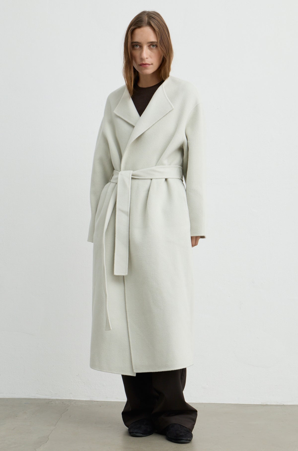 Woman wearing a light grey long coat with a tie belt, brown pants, and a brown t shirt against a light grey wall