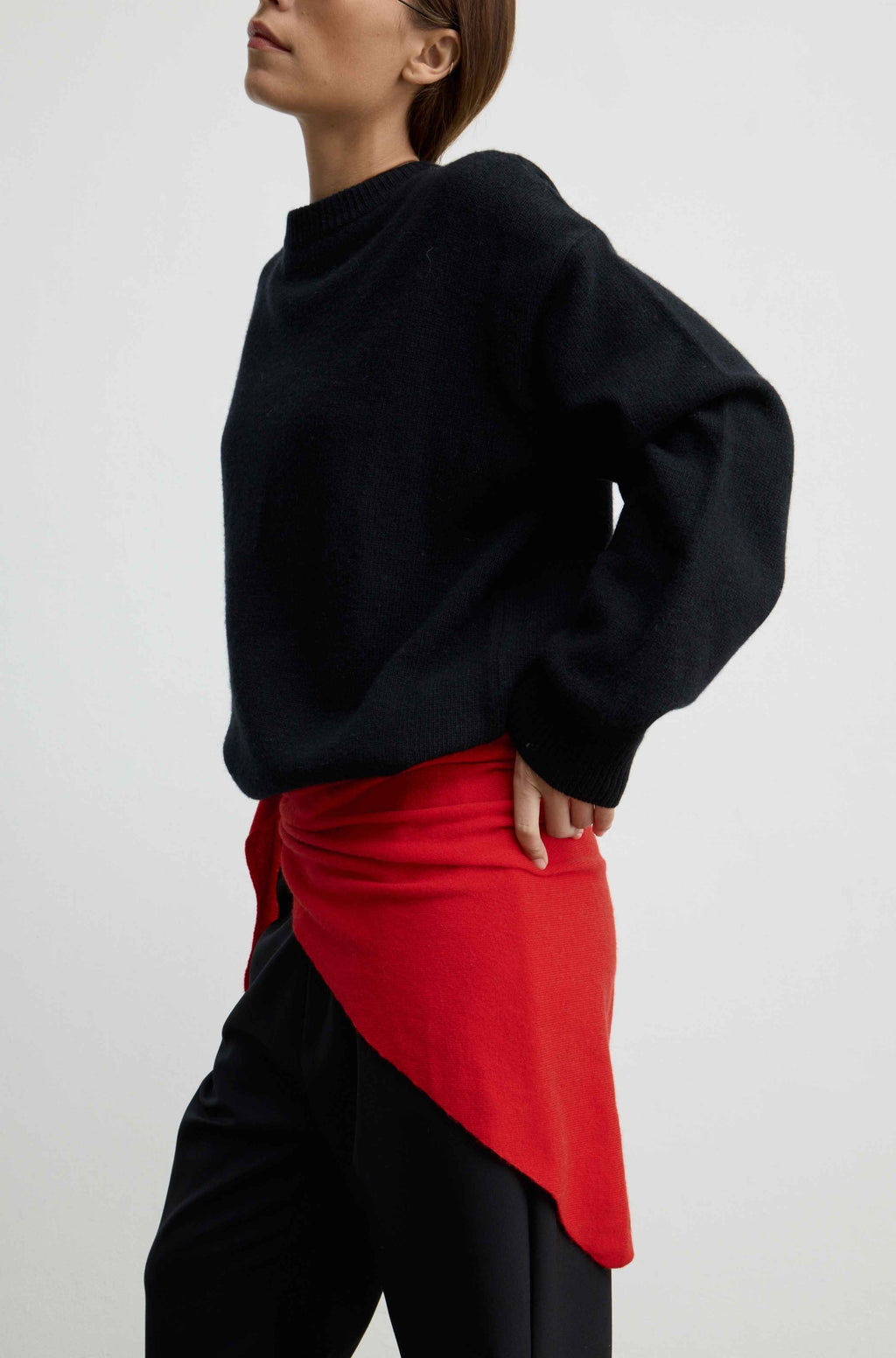 A model wearing a black knit sweater and black pants, styled with a bright red asymmetrical wrap skirt or sash tied at the waist, photographed against a light gray studio background.