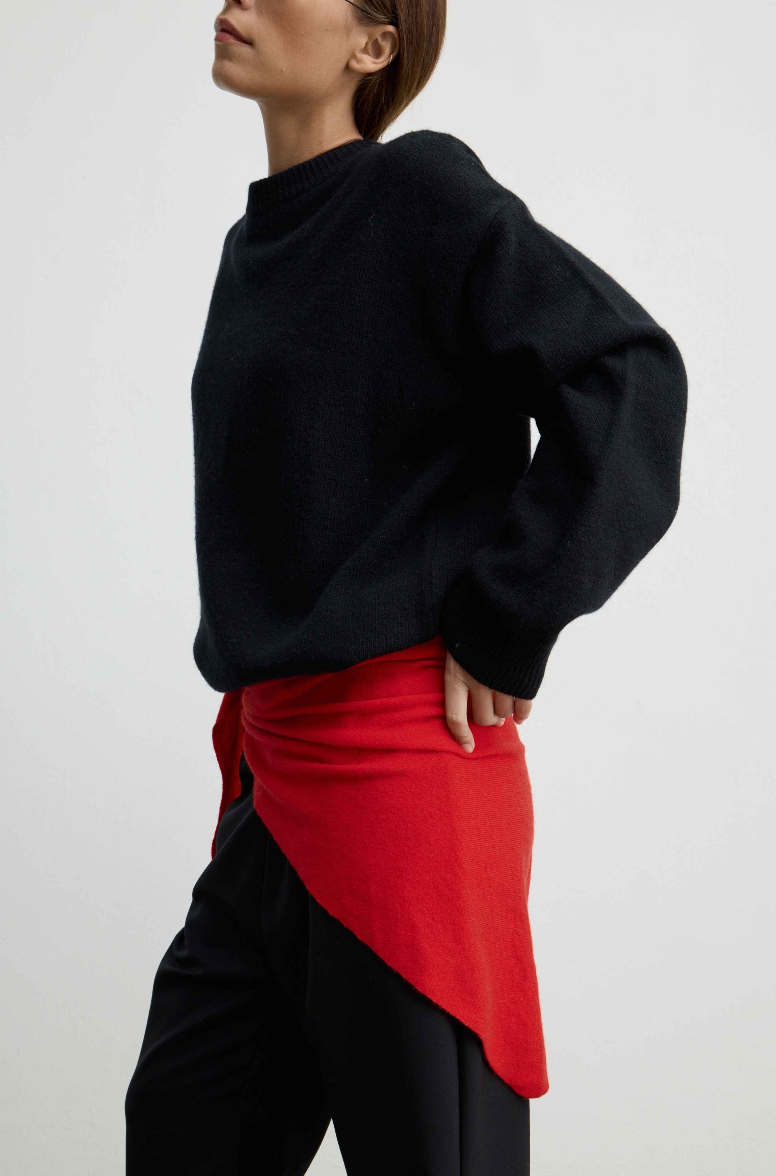 A model wearing a black knit sweater and black pants, styled with a bright red asymmetrical wrap skirt or sash tied at the waist, photographed against a light gray studio background.
